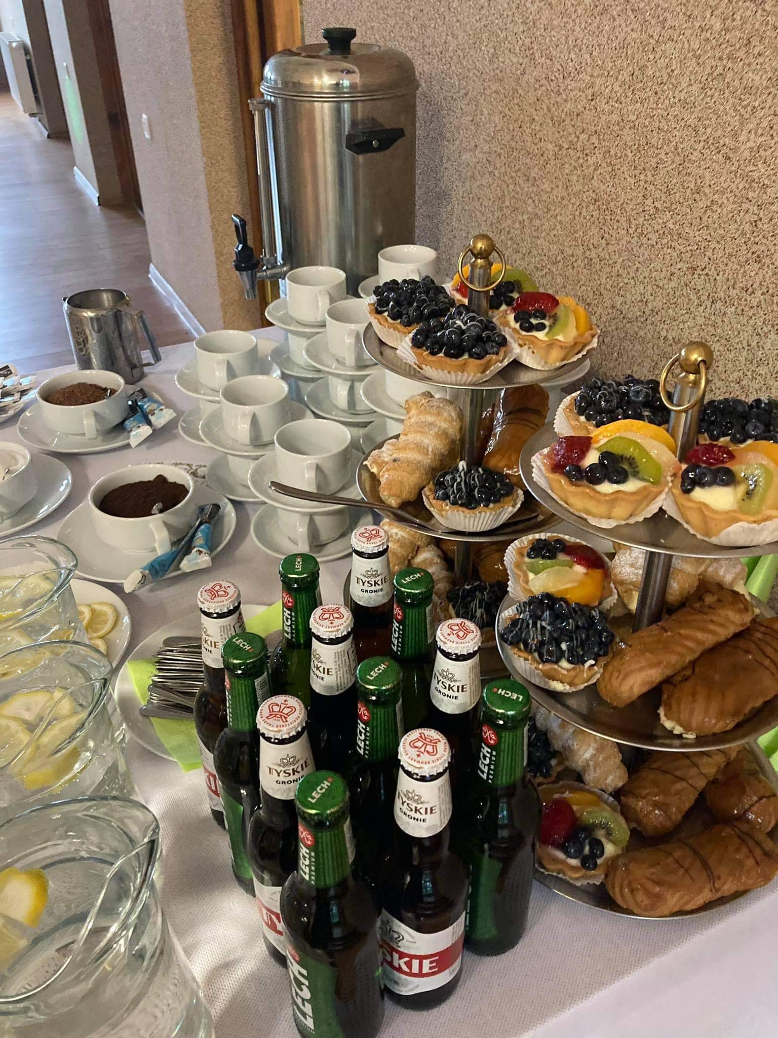 Stół z napojami i przekąskami: dzbanki z wodą z cytryną, butelki piwa, filiżanki z kawą i ciastka z owocami na etażerce.