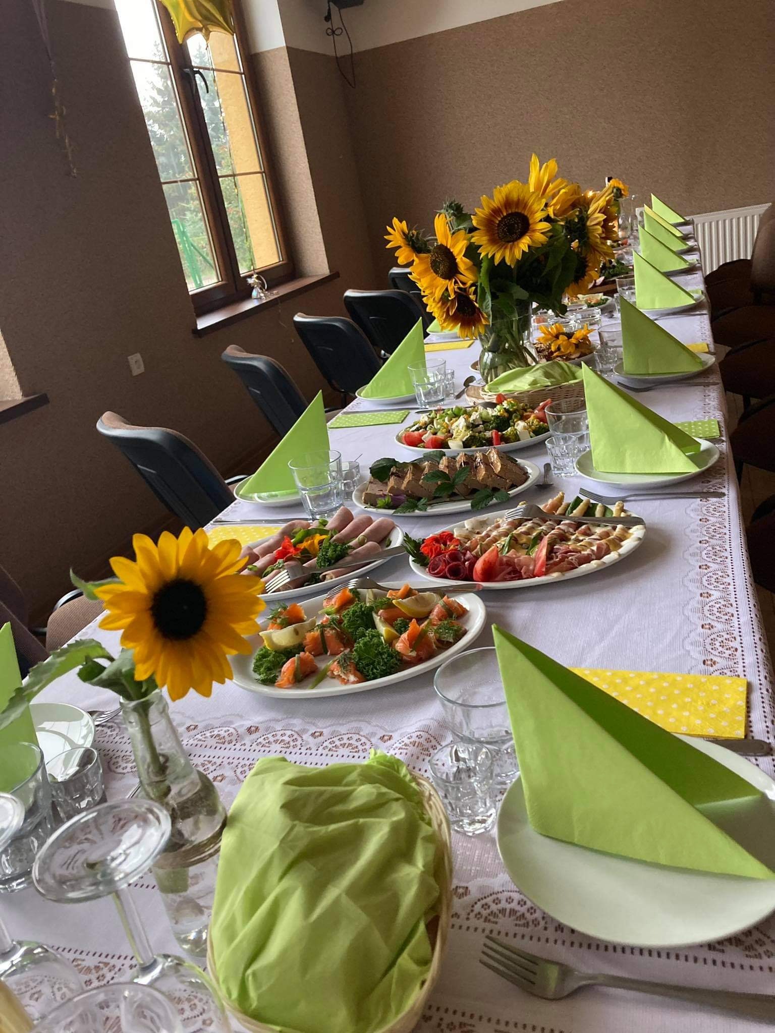 Uroczysty stół nakryty białym obrusem z koronkowym wzorem, dekorowany bukietem słoneczników i pojedynczym słonecznikiem w butelce po napoju, zastawiony talerzami z przekąskami, zielonymi serwetkami...