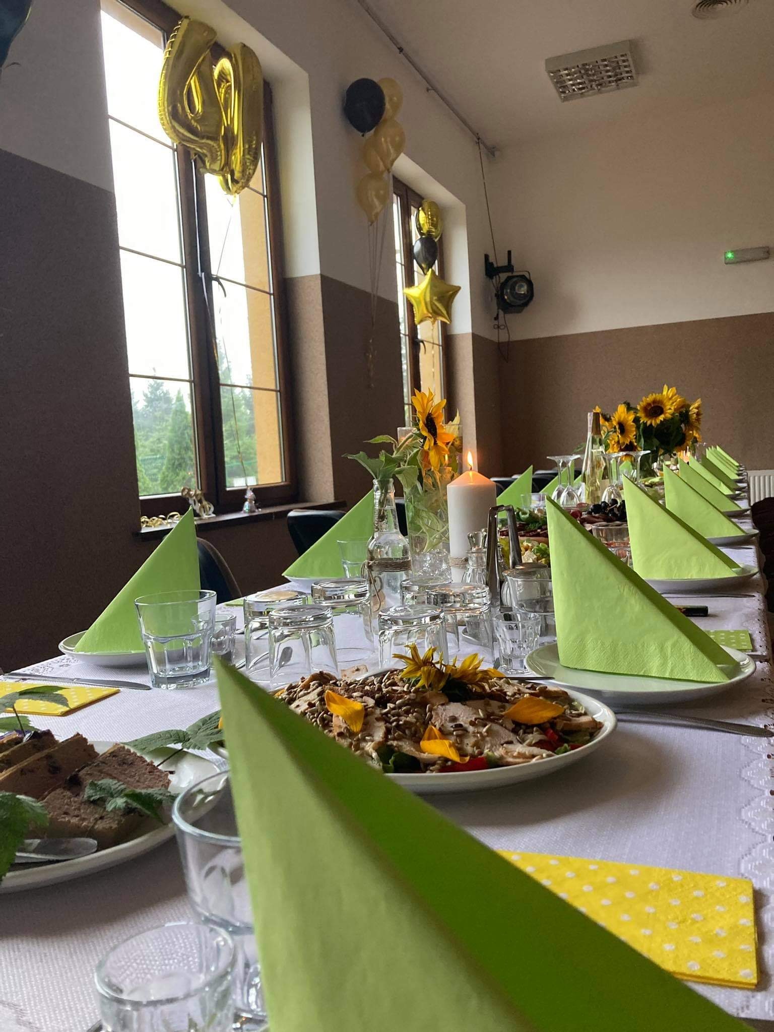 Uroczysty stół nakryty białym obrusem, dekorowany zielonymi serwetkami, kwiatami słonecznika i świecą, z balonami w tle, przygotowany na przyjęcie.