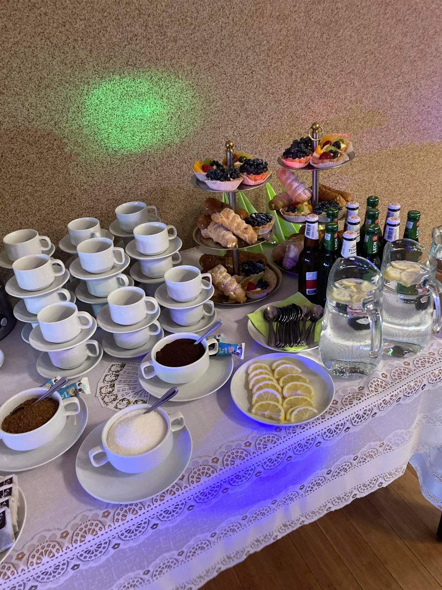 Stół cateringowy z piramidą białych filiżanek, paterą z ciastkami, napojami w szklanych dzbankach z cytryną, oraz dodatkami do kawy na białym obrusie z koronkowym wykończeniem, oświetlony zielonym...
