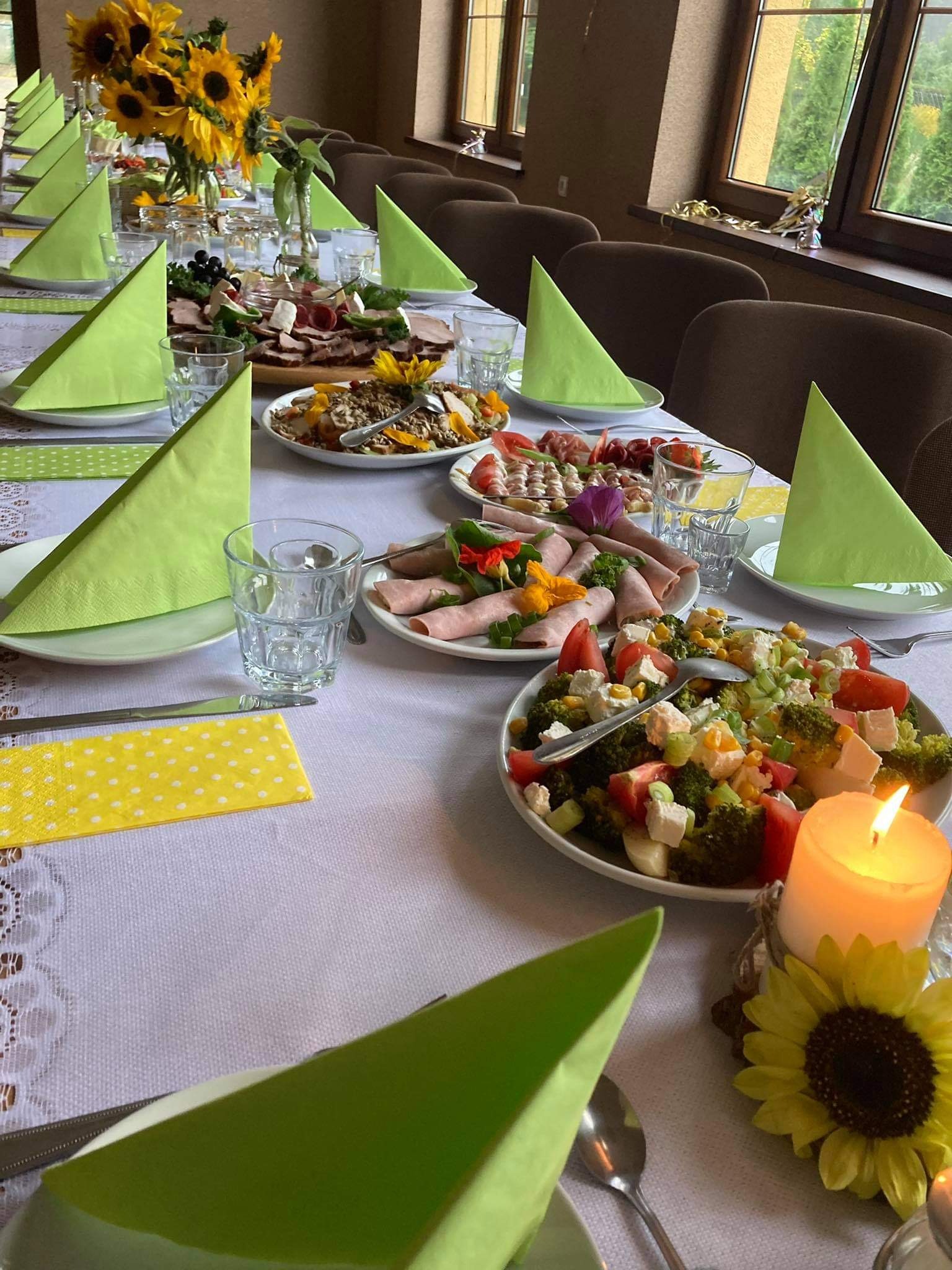 Uroczysty stół z białym obrusem, zielonymi serwetkami w trójkąty, szklankami, talerzami z sałatkami, wędlinami i bukietem słoneczników w wazonie, oświetlony świecą.