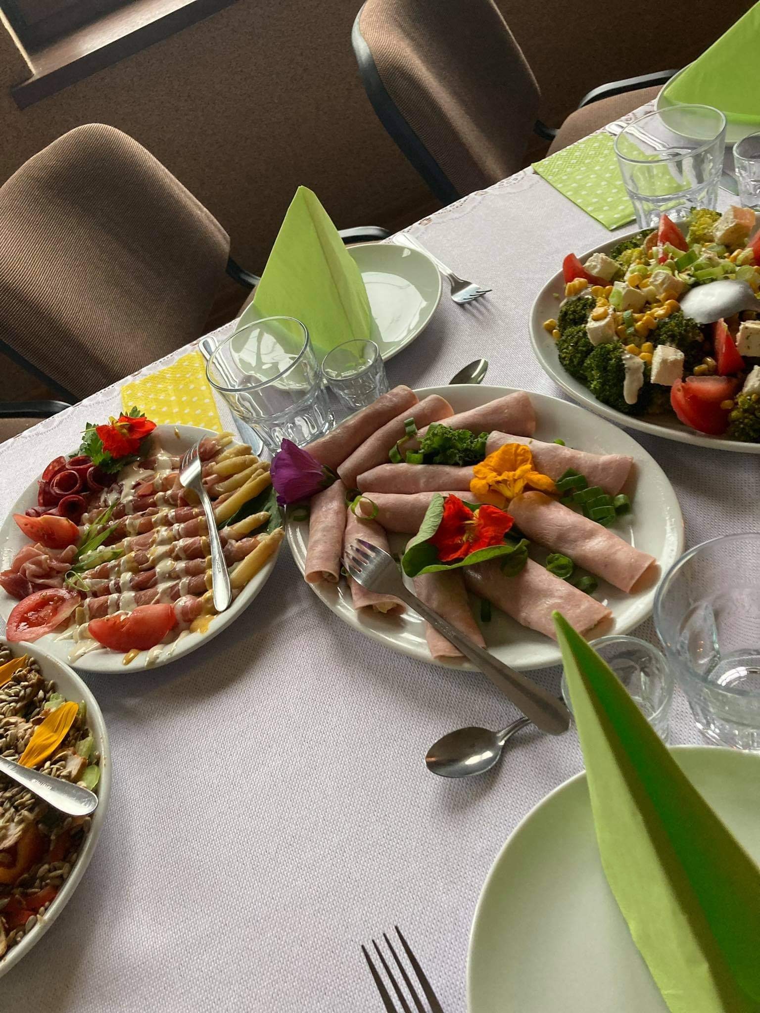 Stół nakryty białym obrusem z aranżacją talerzy z wędlinami, sałatkami i kwiatami, zielone serwetki ułożone w stożki, widoczne sztućce i szklanki.