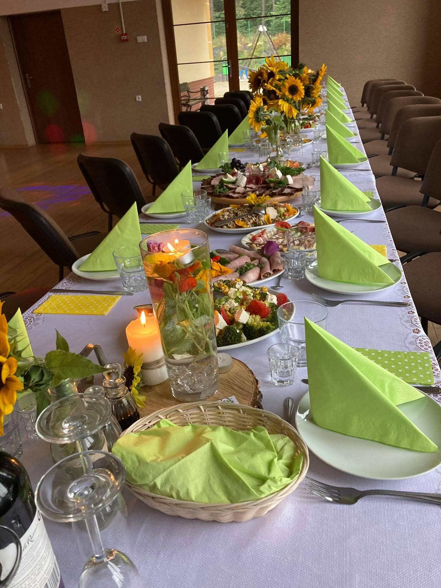 Długi stół z białym obrusem, nakryty dla wielu gości, z zielonymi serwetkami, dekoracjami z żywych słoneczników, świecami i szklanymi naczyniami z kwiatami, zastawiony talerzami z różnorodnymi...