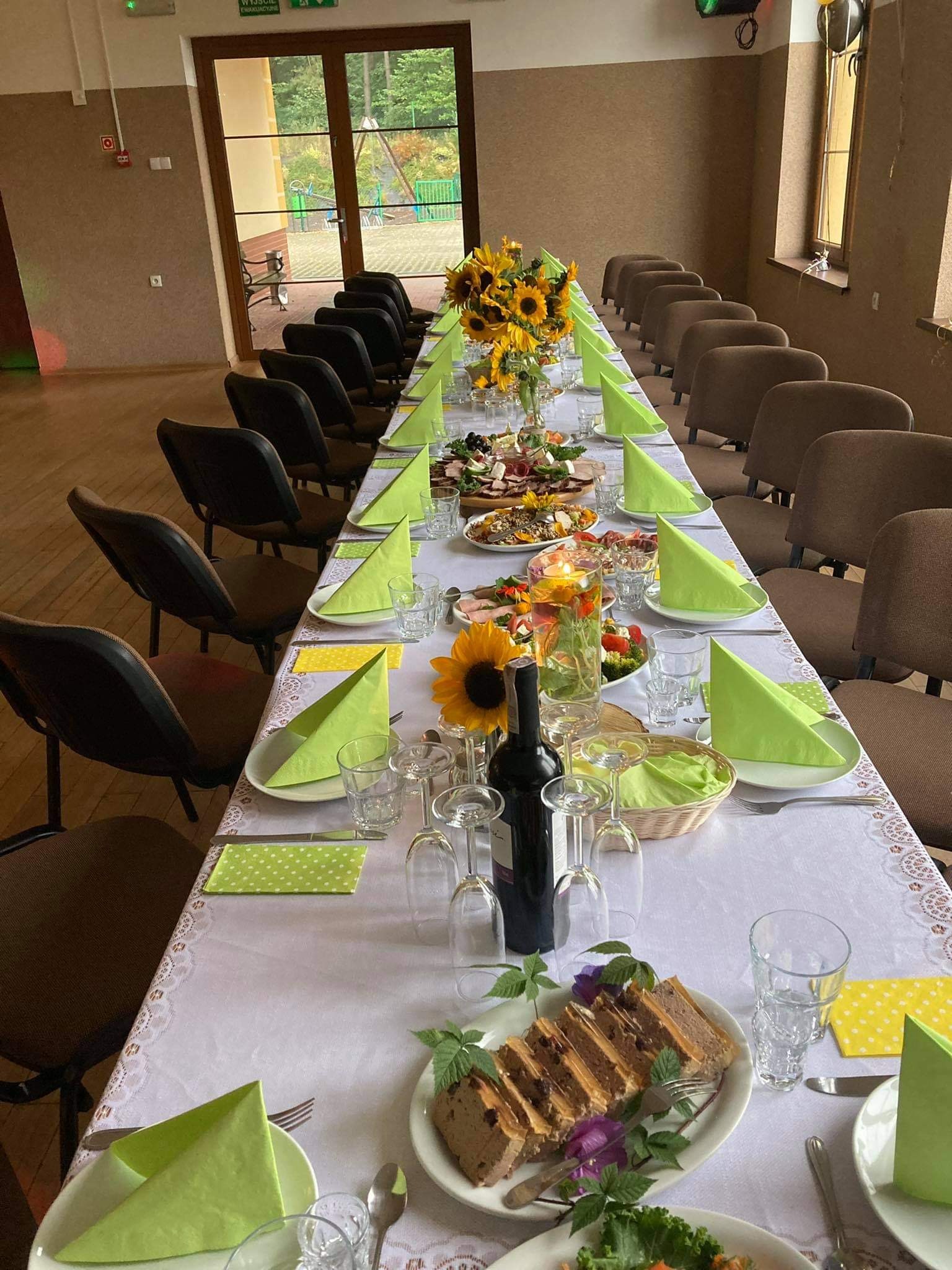 Długi stół nakryty białym obrusem z koronkowym wykończeniem, zastawiony talerzami, szklankami, sztućcami i dekoracjami z zielonych serwetek w kształcie trójkątów oraz wazonów ze słonecznikami.