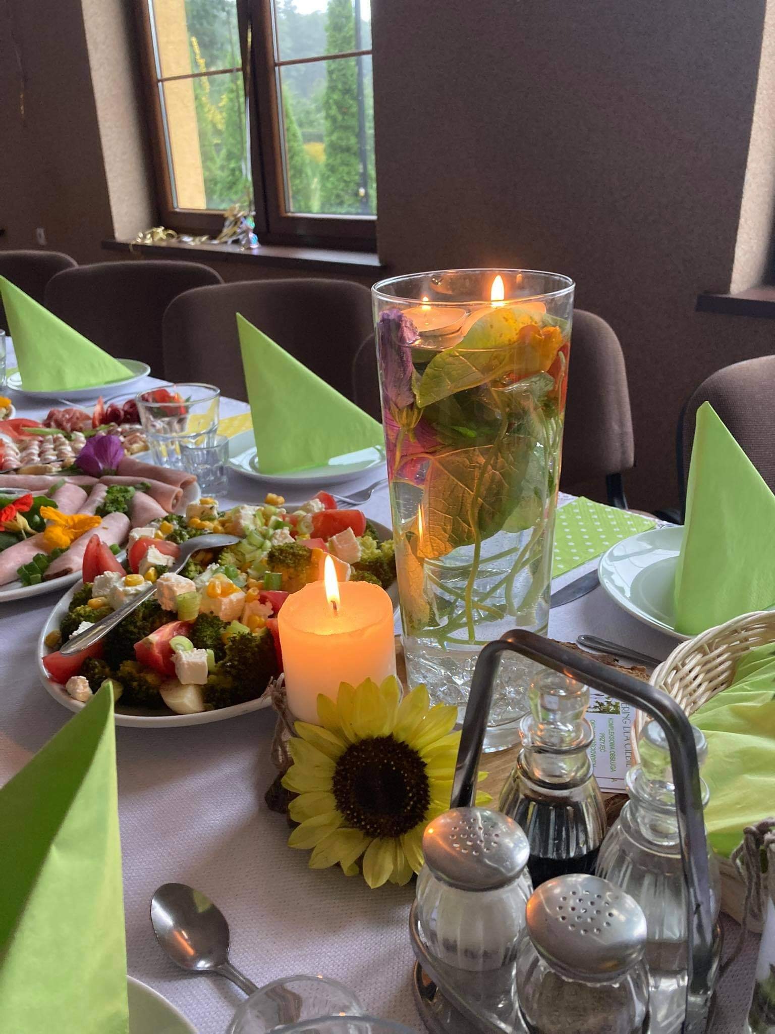 Udekorowany stół z sałatkami, wazonem z kwiatami zatopionymi w wodzie i świecami, nakryty zielonymi serwetkami i srebrnymi sztućcami, w tle okno z widokiem na zieleń.