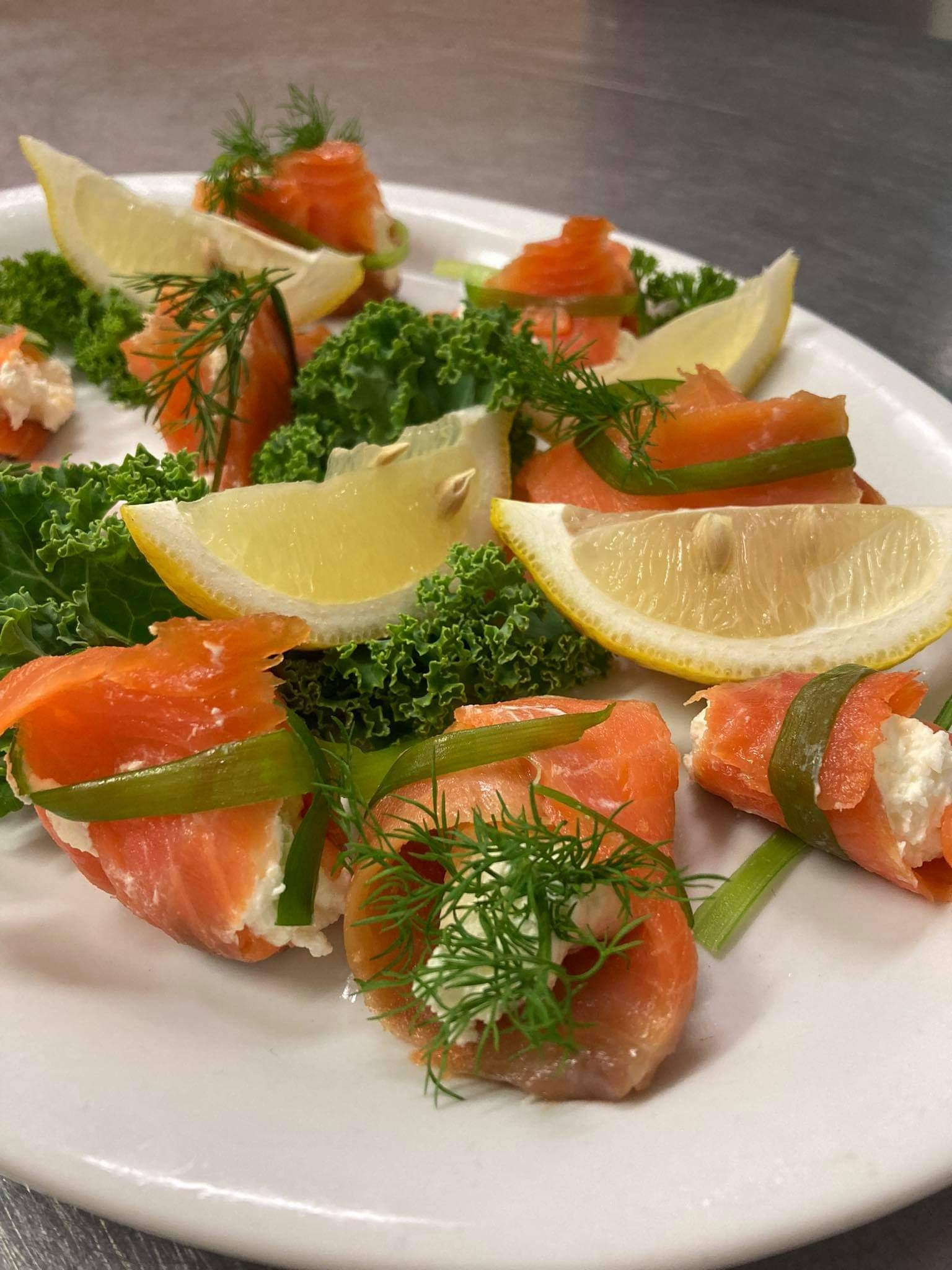 Roladki z łososia z kremowym serem i szczypiorkiem, udekorowane koperkiem, plasterkami cytryny i liśćmi sałaty na białym talerzu.