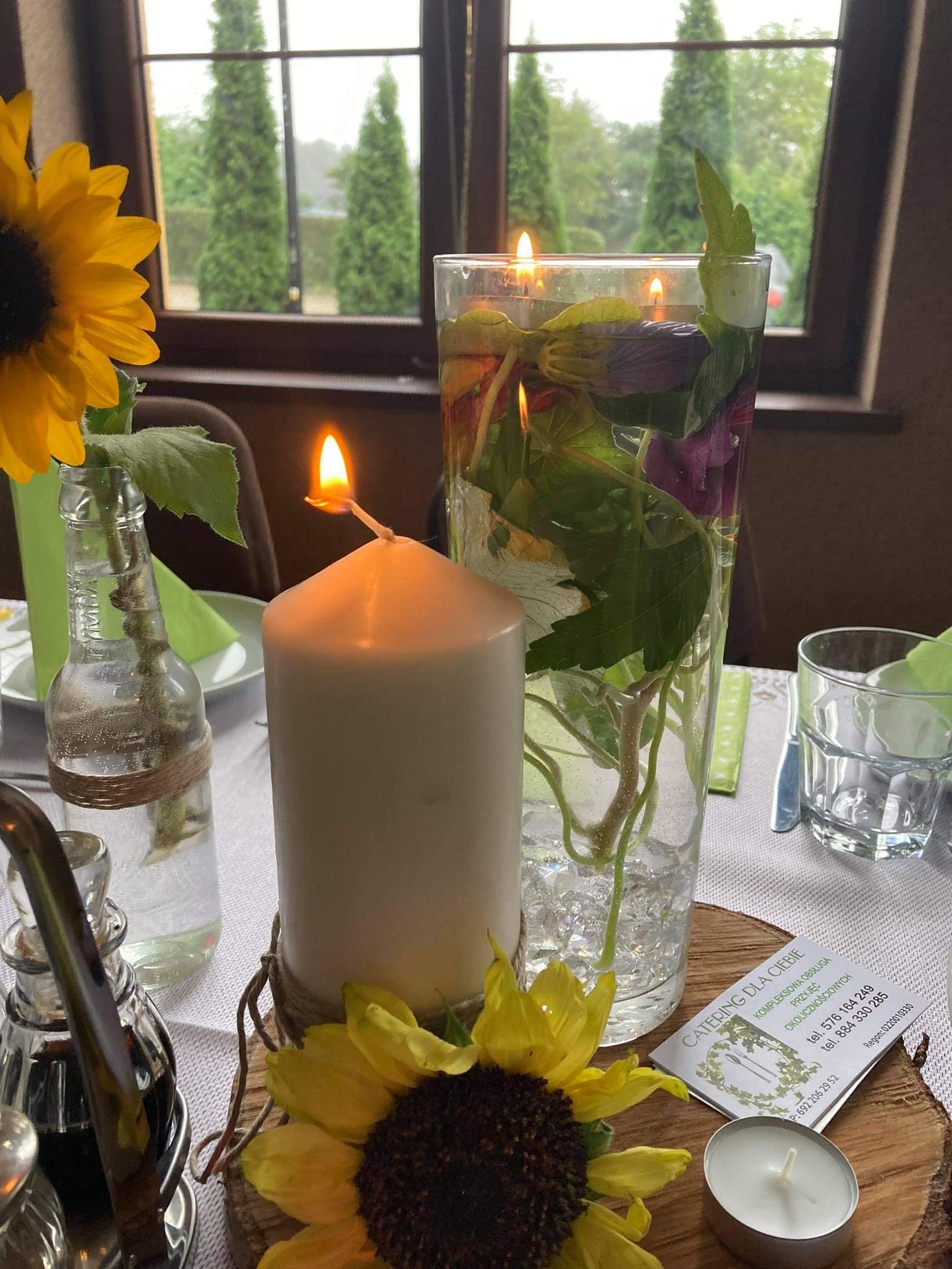 Dekoracja stołu z płonącą świecą, słonecznikami, wazonem z zielonymi liśćmi i wizytówką firmy cateringowej na tle okna z widokiem na drzewa.