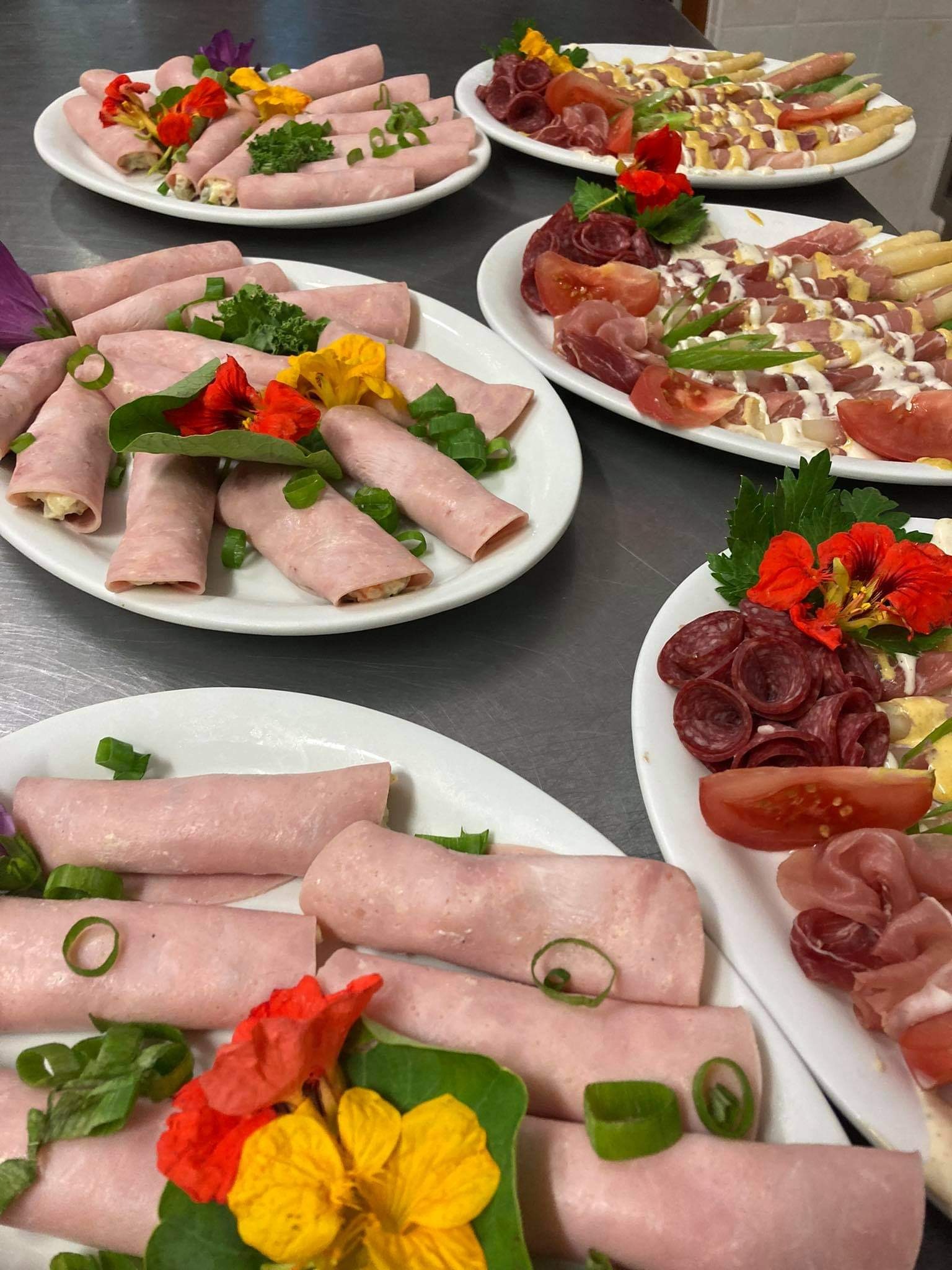 Dekoracyjnie ułożone na białych talerzach roladki z szynki z nadzieniem, salami, szparagi, pomidory, udekorowane jadalnymi kwiatami i szczypiorkiem, na metalowym blacie.