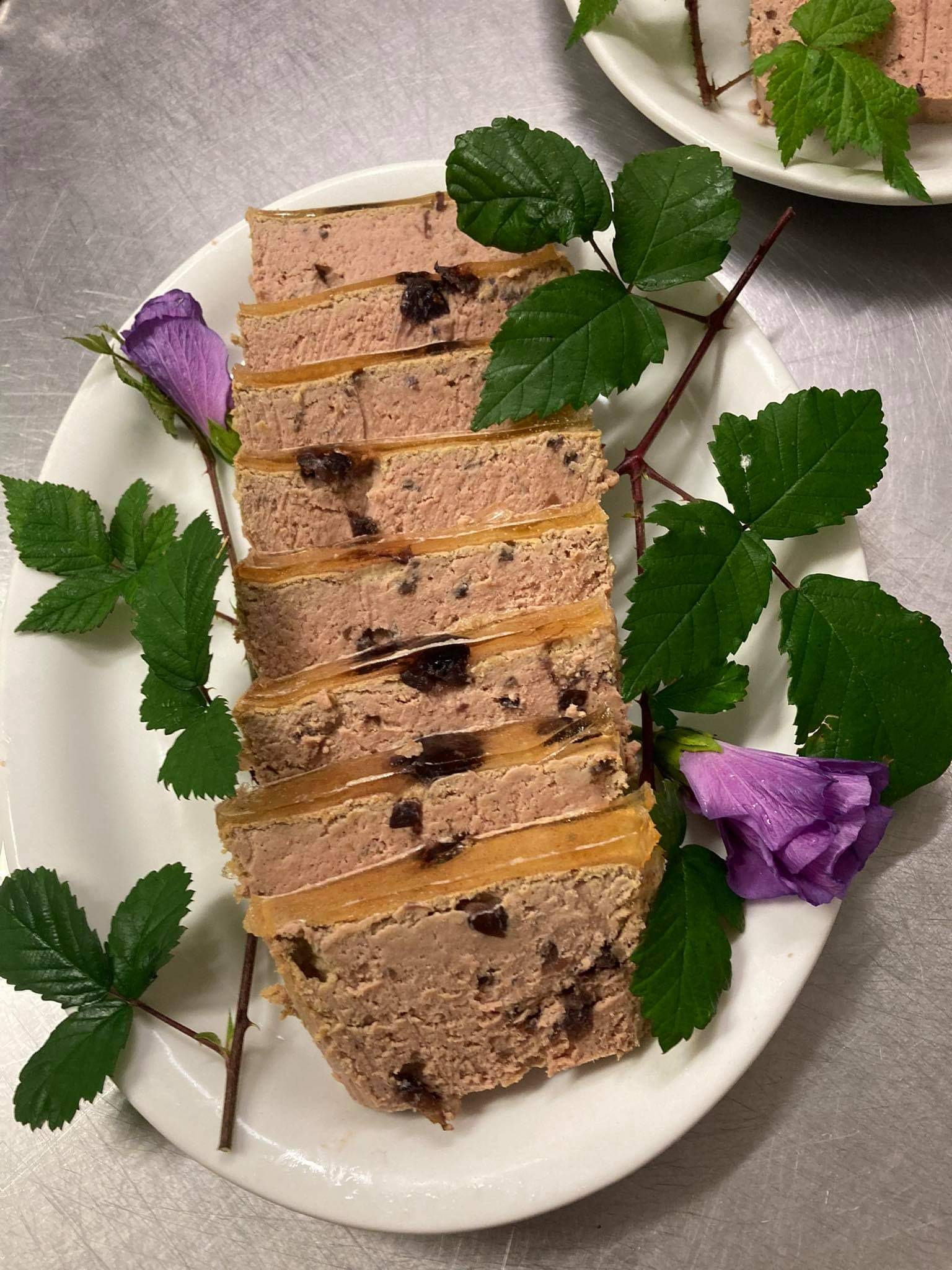 Krojony pasztet z widocznymi kawałkami mięsa i owoców, udekorowany fioletowymi kwiatami i gałązkami z zielonymi liśćmi na białym talerzu, metalowe tło.