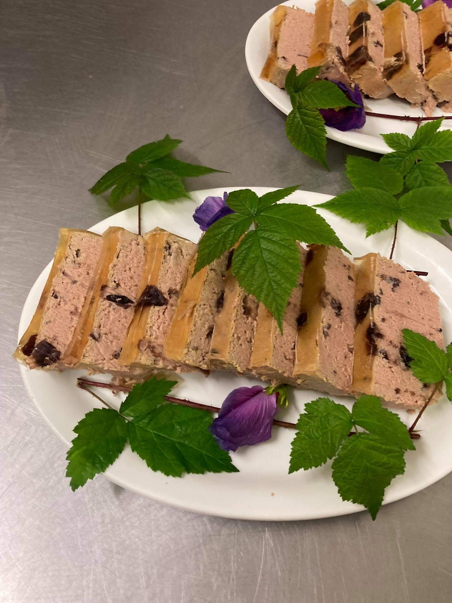 Pasztet z owocami w galarecie, udekorowany liśćmi malin i fioletowymi kwiatami na białym talerzu.