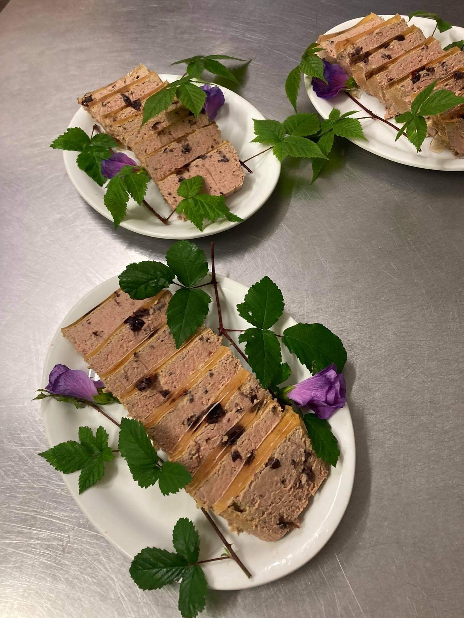Trzy białe talerze z pokrojoną pasztetową, udekorowane zielonymi gałązkami i fioletowymi kwiatami na metalowym blacie.