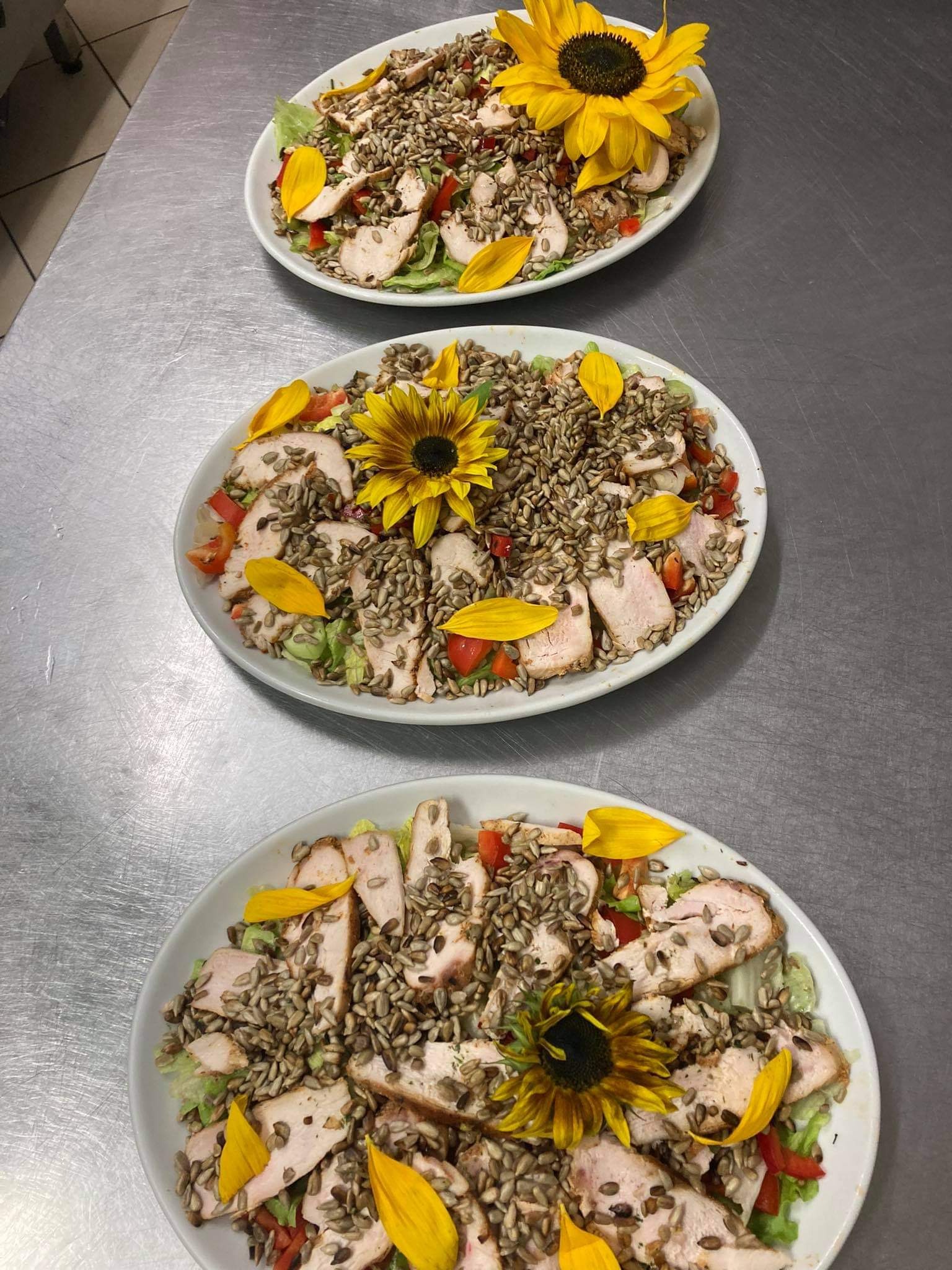 Trzy białe talerze z sałatką z grillowanym kurczakiem, pomidorami, sałatą, pestkami słonecznika i dekoracją z jadalnych płatków słonecznika, ułożone jeden za drugim na metalowym blacie.