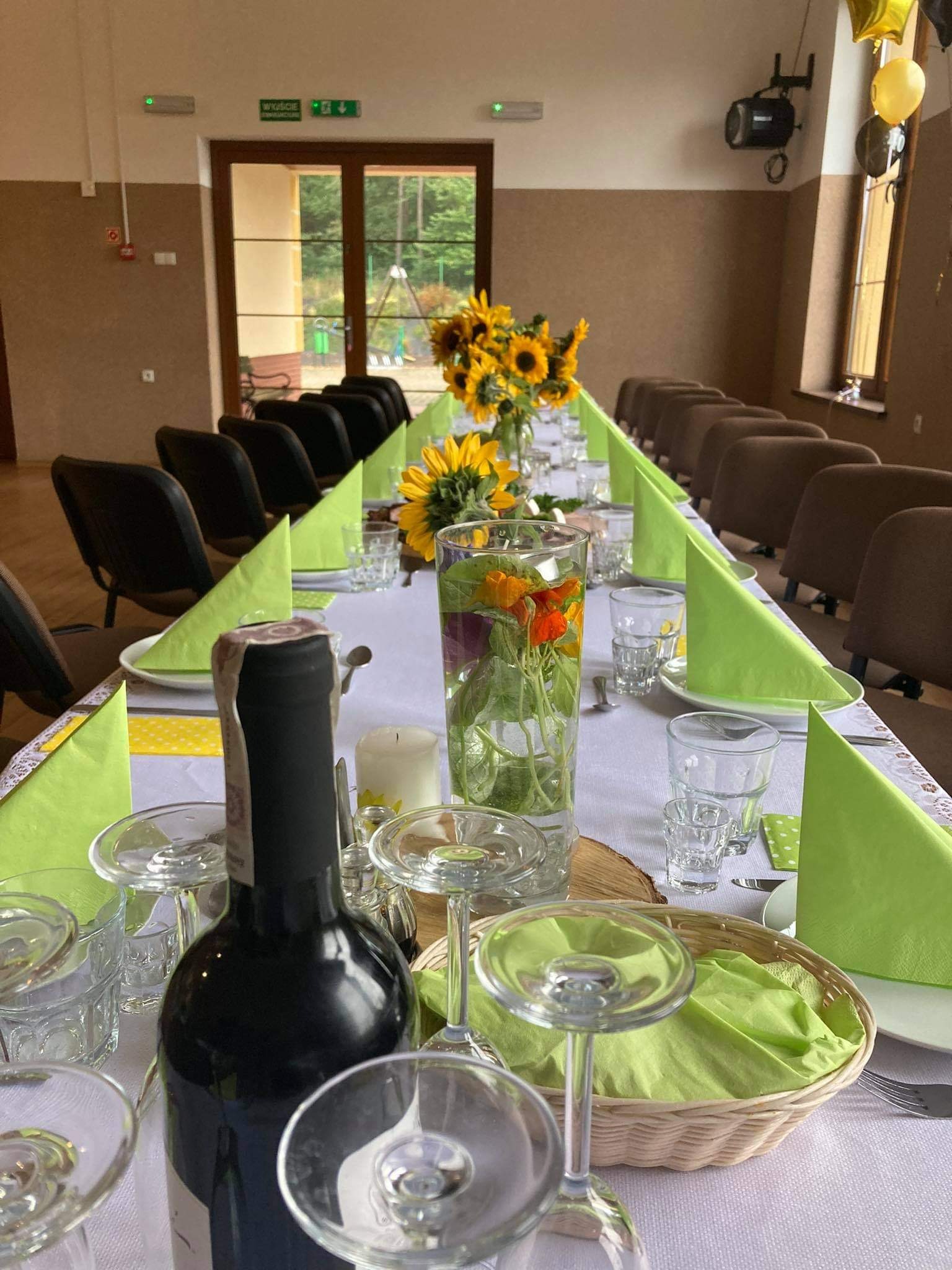 Długi stół nakryty białym obrusem, dekorowany bukietami słoneczników w wazonach, zielonymi serwetkami ułożonymi w trójkąty, szklankami, kieliszkami i butelką wina na pierwszym planie, przygotowany...