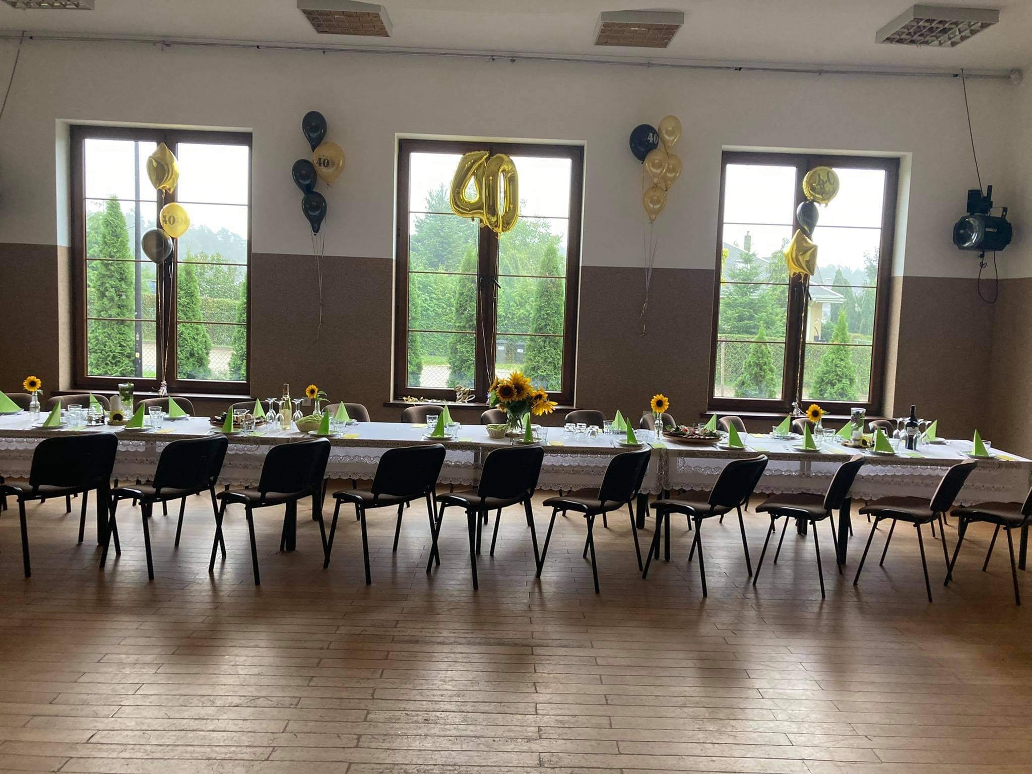 Sala udekorowana na przyjęcie urodzinowe, długi stół z białym obrusem i koronkowym brzegiem, nakryty zielonymi serwetkami, szklankami i kwiatami słonecznika, czarne krzesła, okna z balonami...