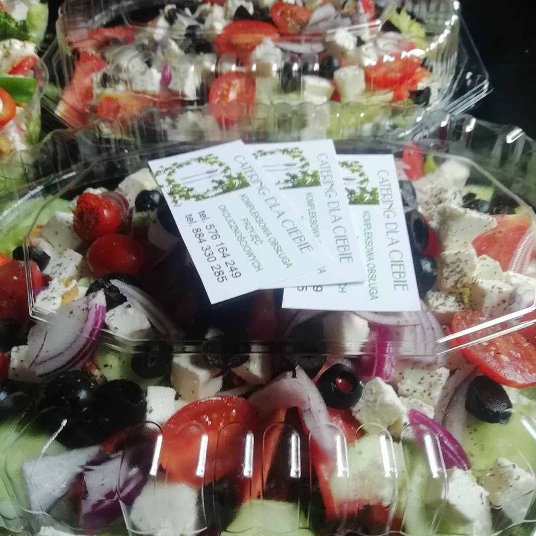 Sałatka grecka w plastikowym opakowaniu z widocznymi składnikami: pomidory, ogórki, oliwki, czerwona cebula i ser feta, uzupełniona o wizytówki firmy cateringowej.