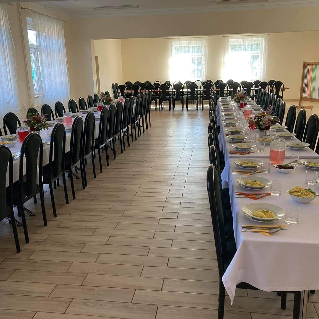 Długie stoły z białymi obrusami i czarnymi krzesłami, nakryte talerzami z makaronem, szklankami i dzbankami z napojem, ustawione w dużej sali z jasnymi ścianami i oknami z białymi firankami...