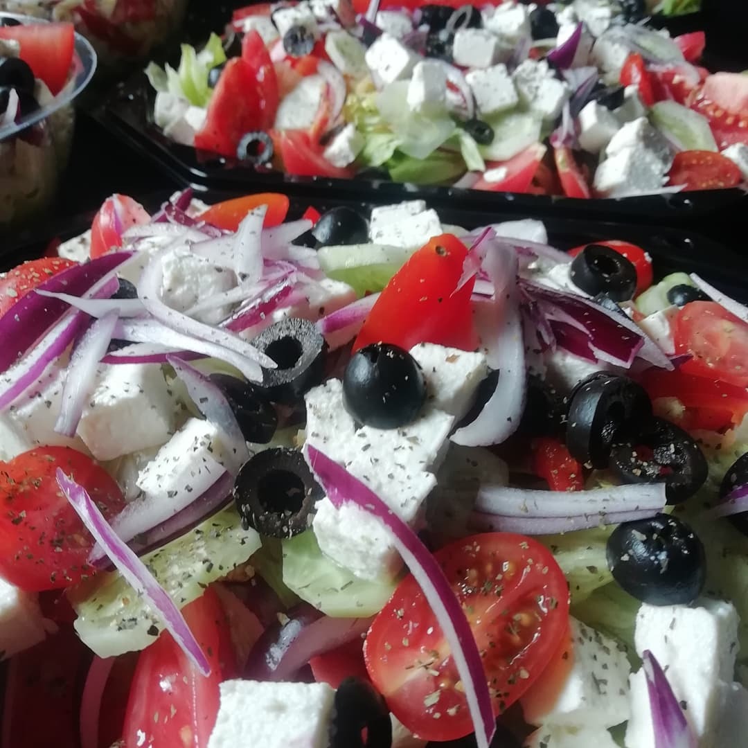 Dwa czarne pojemniki z sałatką grecką: kostki sera feta, pomidory, ogórki, czerwona cebula i oliwki. Pierwszy pojemnik jest w ostrości, drugi w tle.