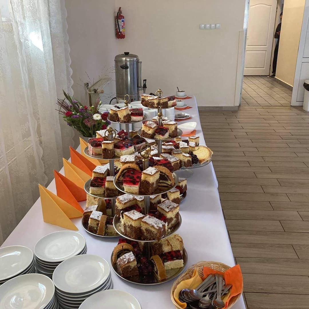 Stół cateringowy z wielopoziomowymi paterami pełnymi różnorodnych ciast, obok stos białych talerzy, pomarańczowe serwetki, kwiaty, termos i koszyk ze sztućcami