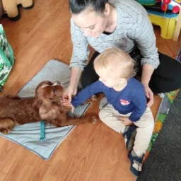 Kobieta z dzieckiem głaszczą brązowego psa rasy Cavalier King Charles Spaniel leżącego na niebieskiej podkładce w pokoju zabaw.