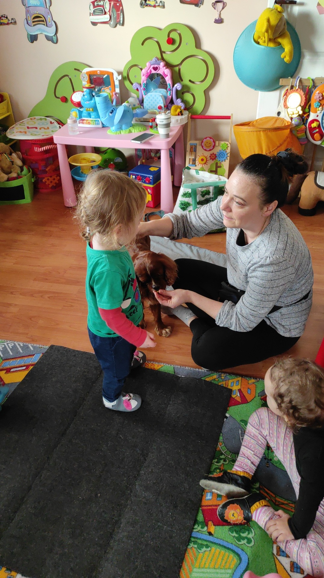 Wnętrze pokoju zabaw z dziećmi i psem rasy Cavalier King Charles Spaniel, karmionym z ręki przez opiekunkę, na podłodze kolorowy dywan i czarna mata, w tle zabawki i mebelki dla dzieci.