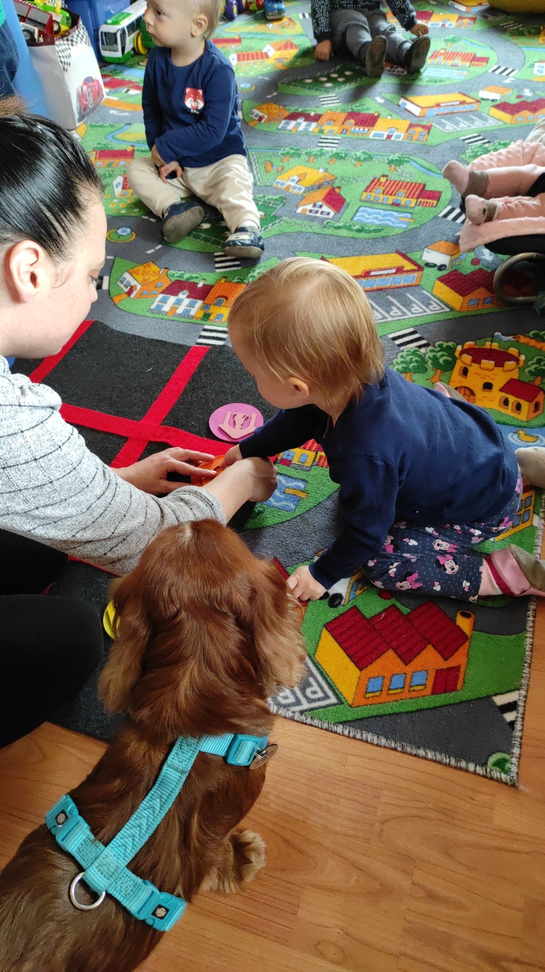 Dzieci bawią się z opiekunką na dywanie z motywem miasta, w pobliżu siedzi pies w niebieskiej uprzęży.