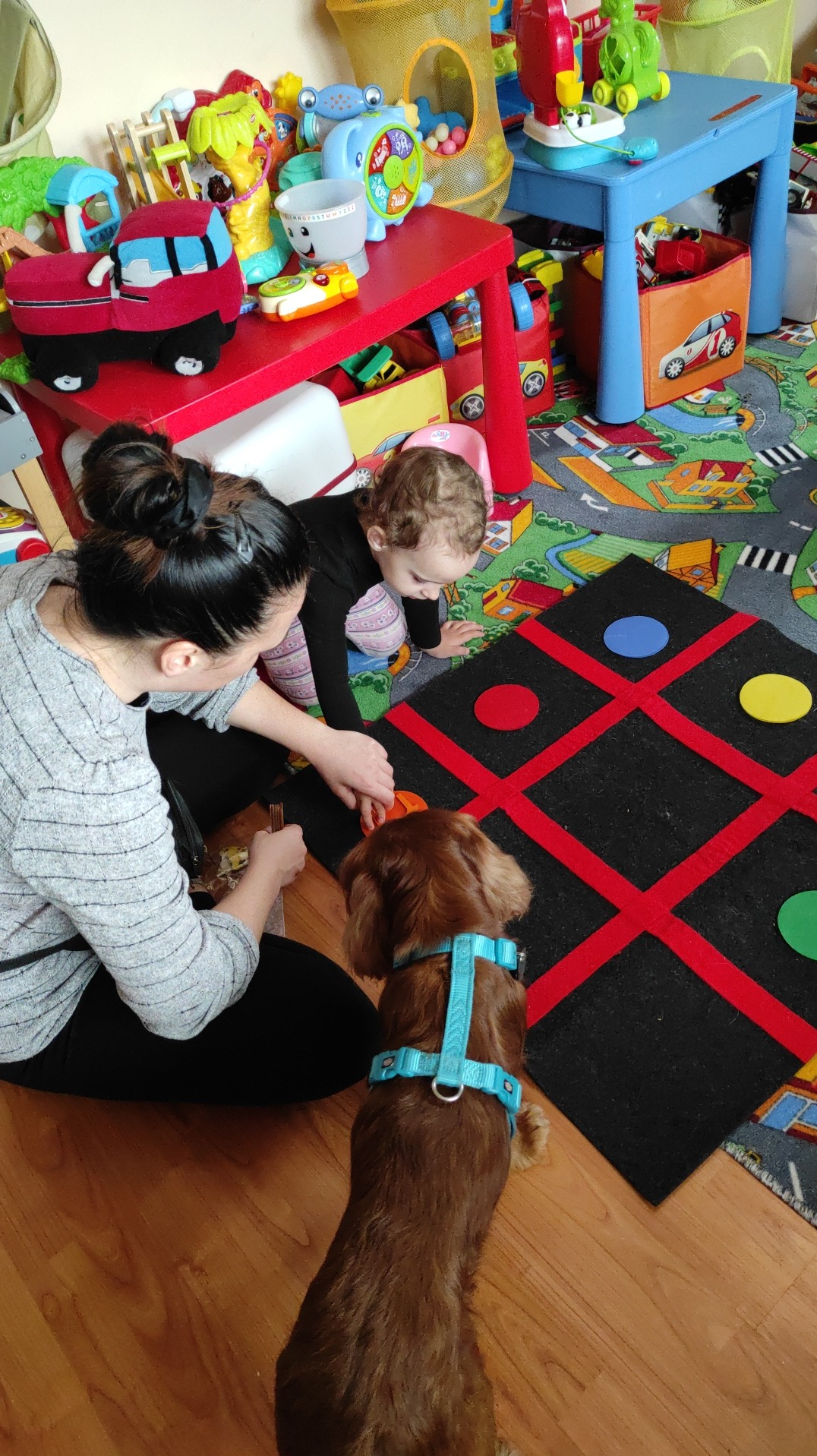 Kobieta z dzieckiem grają w kółko i krzyżyk na dużej macie edukacyjnej, obok siedzi pies w niebieskich szelkach, w tle kolorowe zabawki i meble dziecięce.