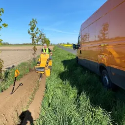 Operator na żółtej maszynie wykopuje wąski rów w ziemi, obok stoi pomarańczowy van, w tle pole i niebieskie niebo.