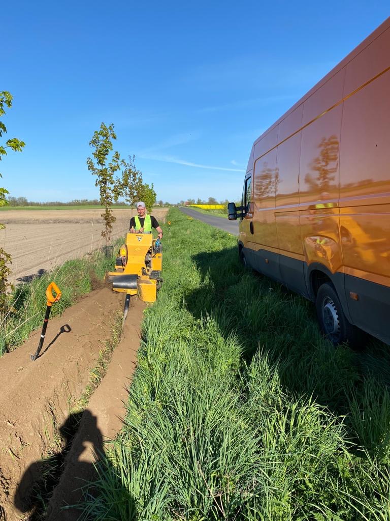 Operator na żółtej maszynie wykopuje wąski rów w ziemi, obok stoi pomarańczowy van, w tle pole i niebieskie niebo.