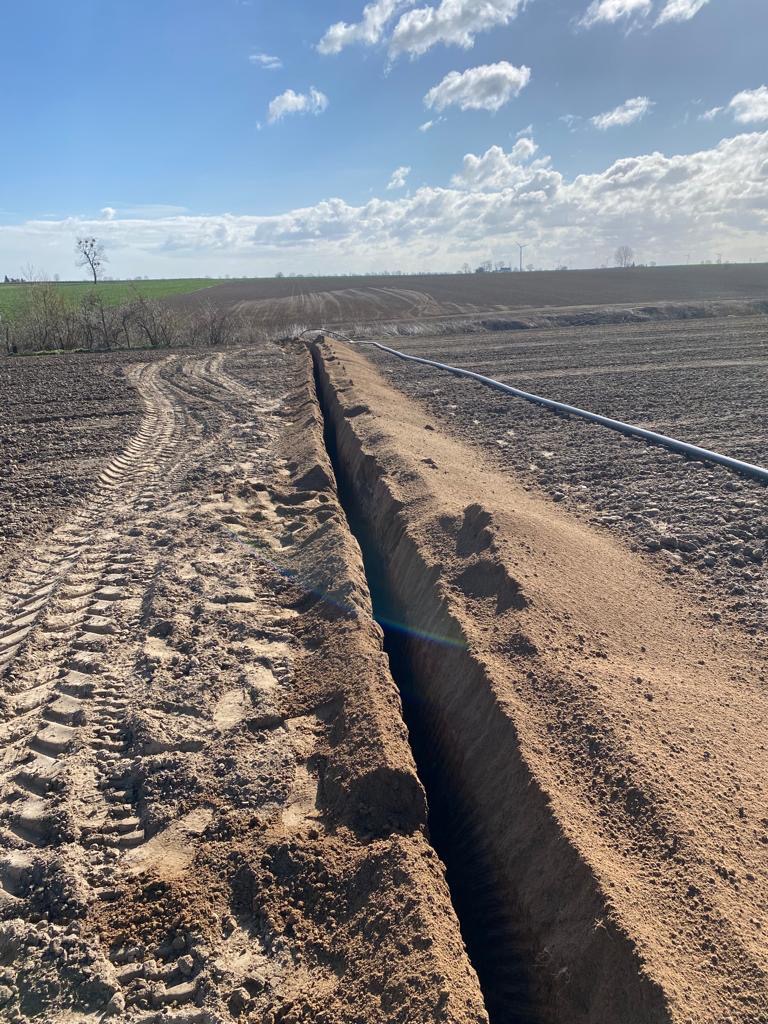 Świeżo wykopany, głęboki rów w ziemi uprawnej z widocznymi śladami opon ciągnika i rurą na powierzchni, w tle pole i błękitne niebo z chmurami.