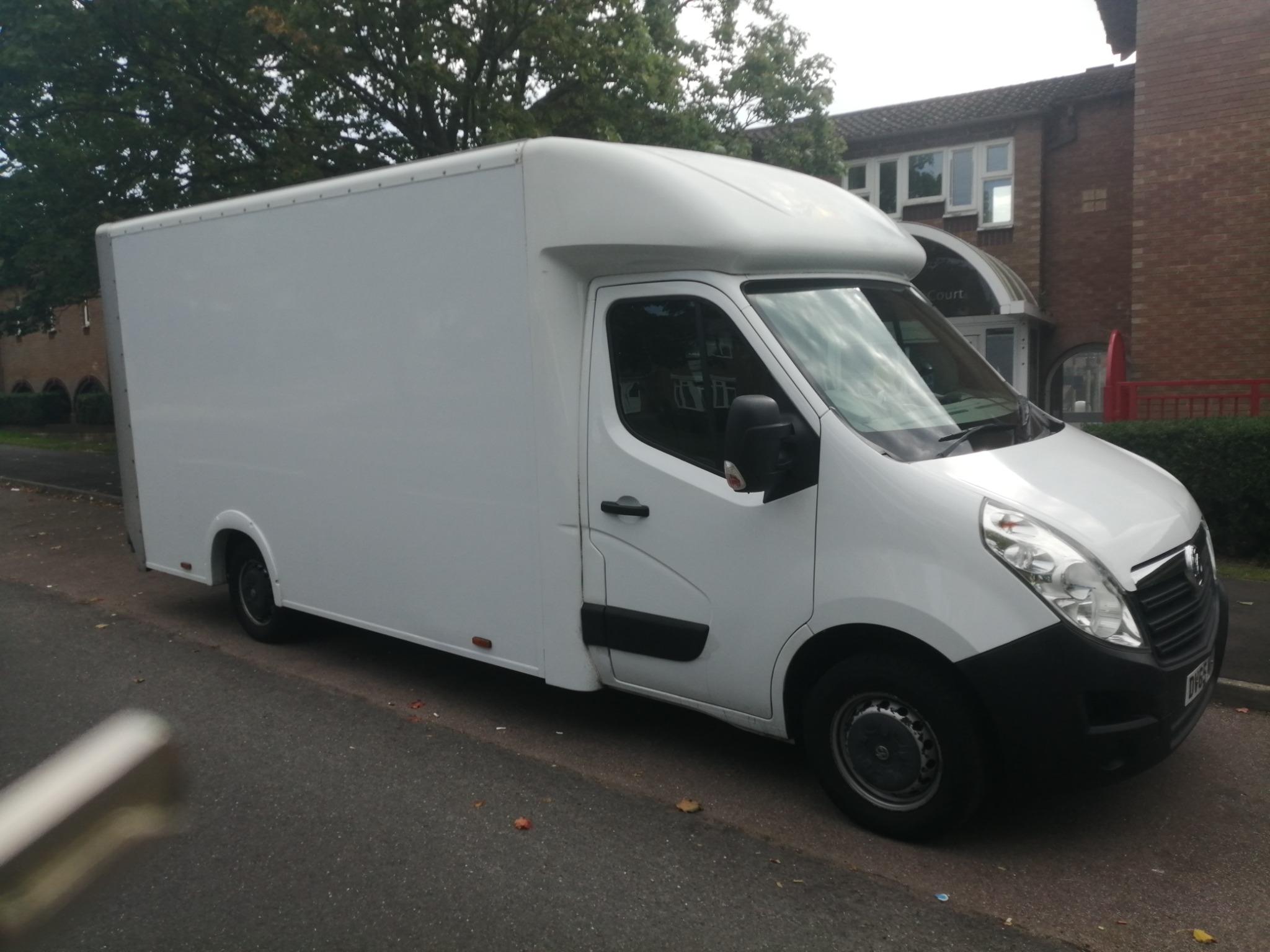 Biały samochód dostawczy typu furgon z wysoką kabiną, zaparkowany na ulicy w pobliżu budynku mieszkalnego z cegły.