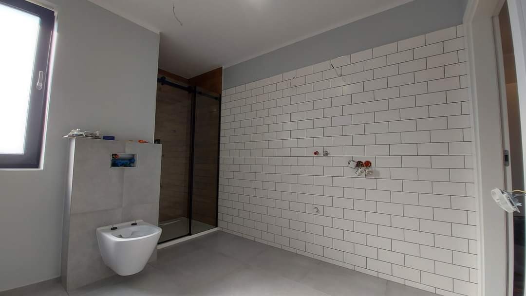 Wnętrze łazienki w trakcie remontu: białe, prostokątne płytki ułożone na ścianie, zamontowany stelaż podtynkowy WC z częściowo widoczną instalacją, kabina prysznicowa z czarnymi profilami.