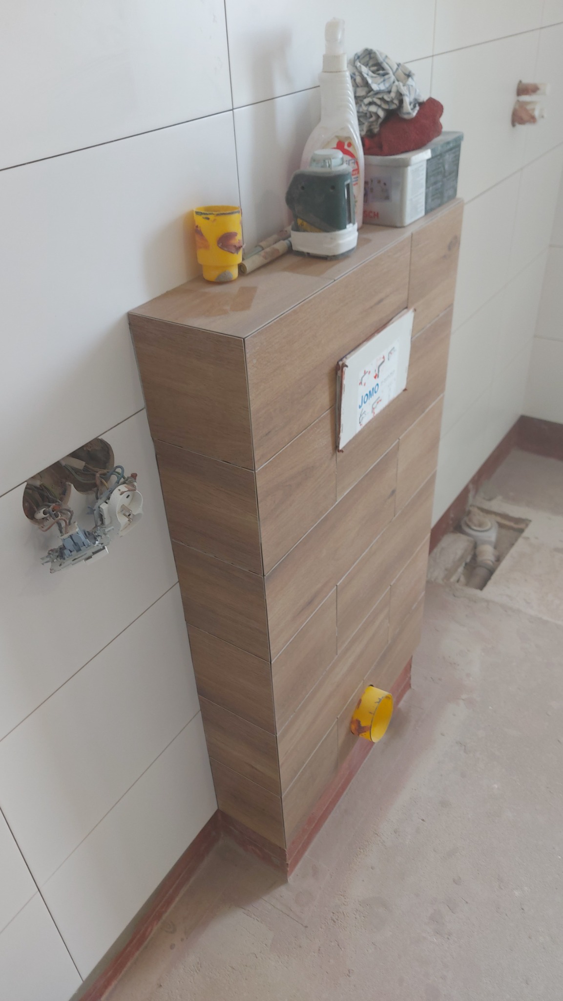 Obudowa stelaża podtynkowego WC wykończona płytkami imitującymi drewno, widoczny otwór kanalizacyjny z żółtą rurą w Knurowie, białe płytki na ścianie oraz narzędzia i chemia na obudowie.