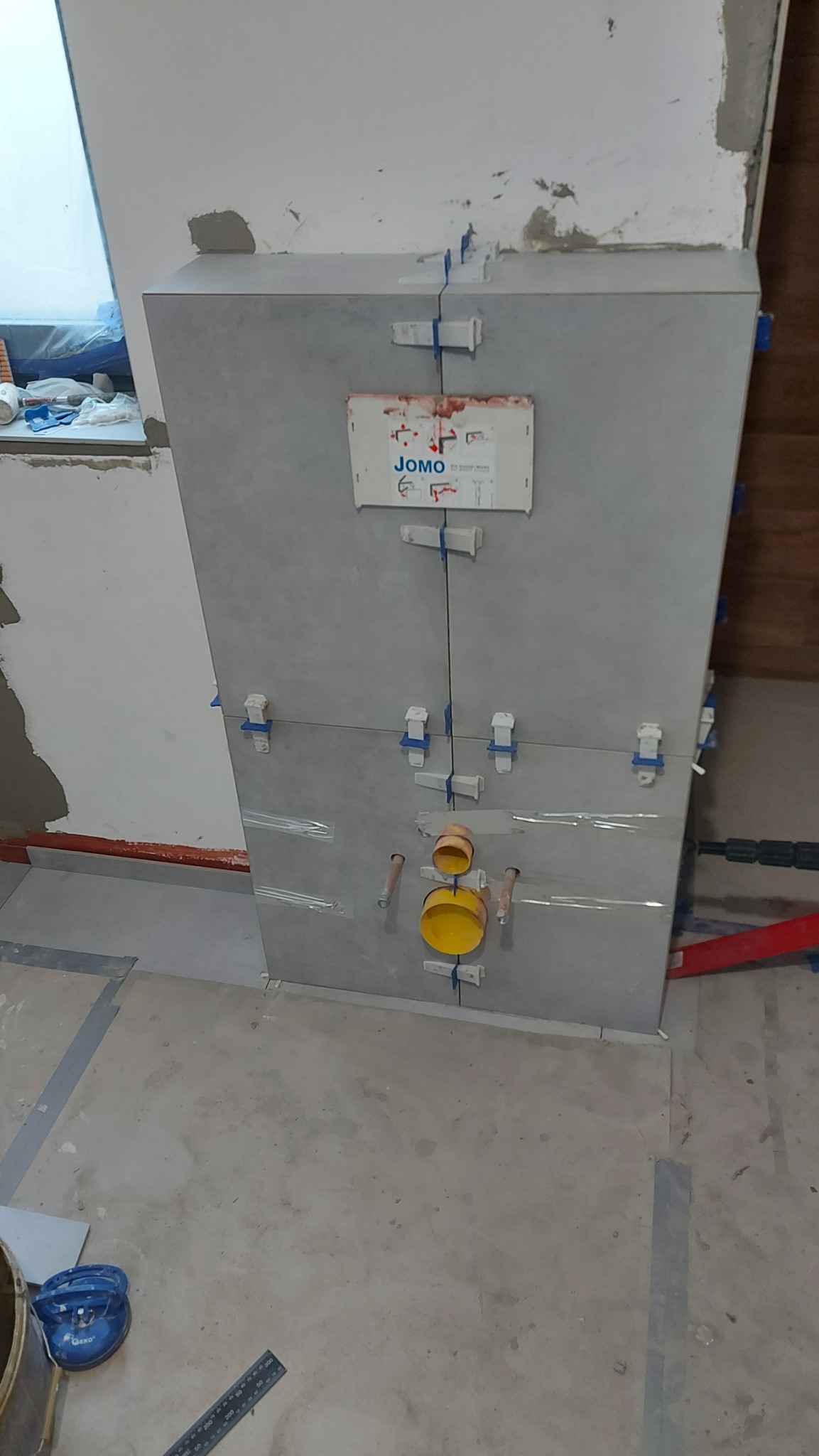 Montaż szarej, ceramicznej obudowy stelaża WC z widocznymi elementami instalacji wodno-kanalizacyjnej, krzyżykami dystansowymi i przyklejoną instrukcją JOMO w trakcie prac wykończeniowych łazienki.