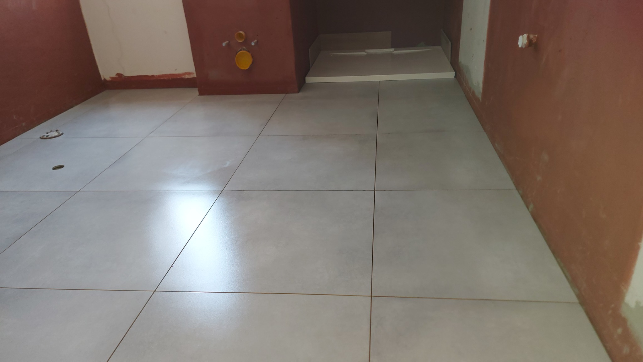 Świeżo ułożone, duże, szare płytki podłogowe w trakcie remontu łazienki, widoczne fragmenty ścian przygotowanych pod malowanie i instalacje sanitarne.