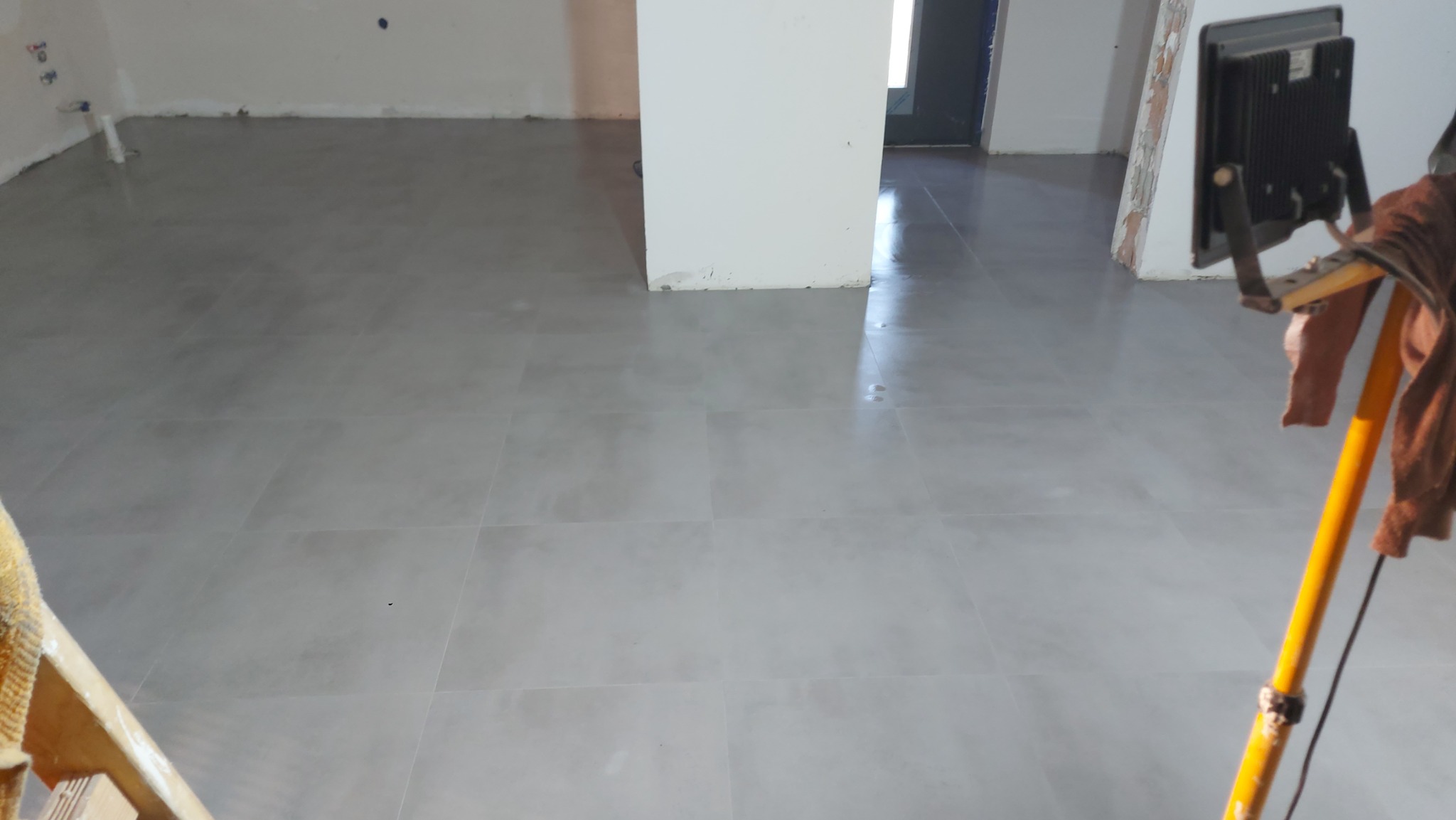 Świeżo ułożone, jasnoszare płytki podłogowe w pustym pomieszczeniu, oświetlone lampą roboczą na żółtym statywie, widoczne ślady po fugowaniu.