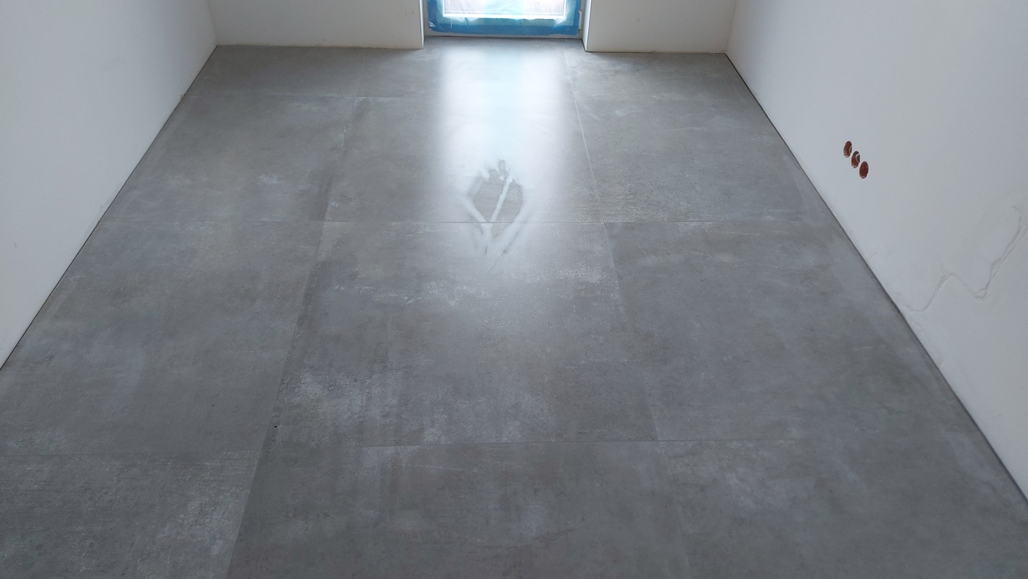 Szare, matowe płytki podłogowe ułożone w nowym pomieszczeniu, widoczne ślady po czyszczeniu w centralnej części, surowe ściany z widocznymi otworami na gniazdka elektryczne.