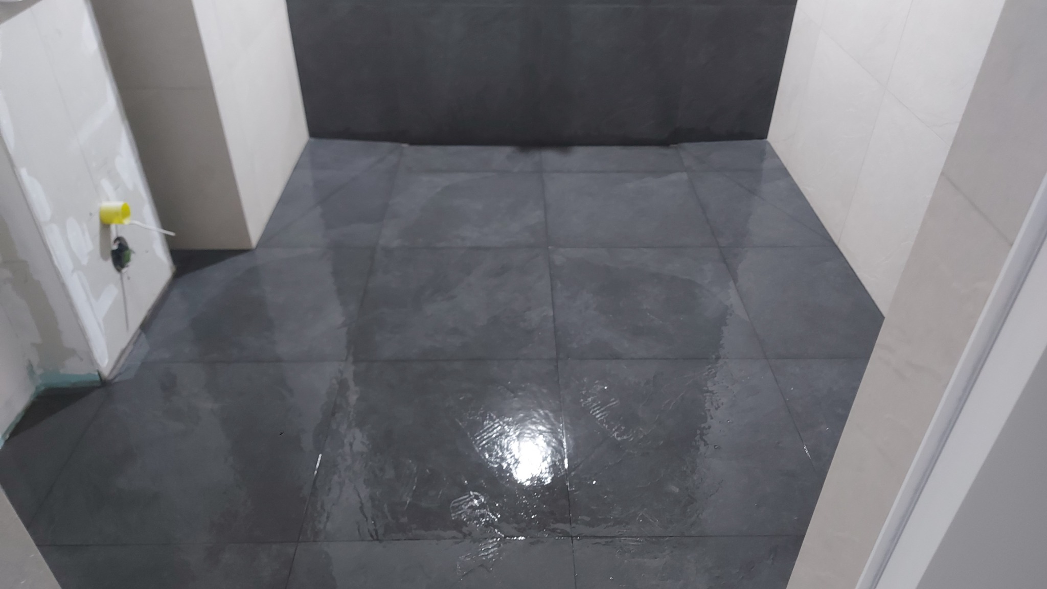 Świeżo położone, ciemnoszare płytki podłogowe w łazience, jeszcze mokre po myciu, z widocznymi fragmentami ścian w trakcie wykończenia i żółtą osłoną instalacji elektrycznej w rogu.