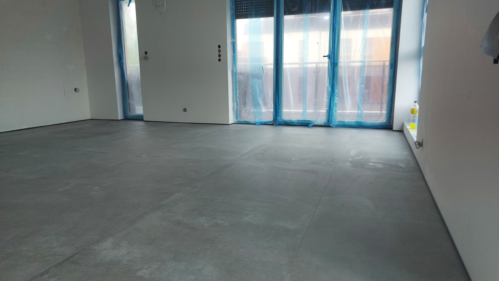 Szare płytki podłogowe w surowym wnętrzu, okna zabezpieczone folią malarską, widoczne puszki elektryczne w ścianie, butelka z płynem na parapecie.