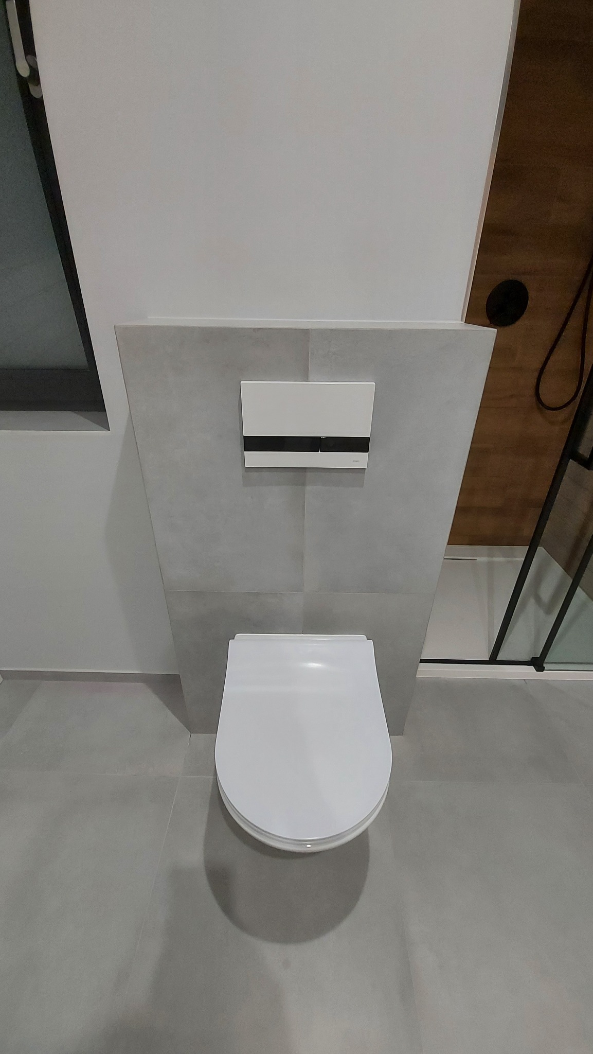 Nowoczesna, podwieszana toaleta z białym sedesem i szarą spłuczką w zabudowie, na tle szarych płytek podłogowych i fragmentu kabiny prysznicowej z czarną ramą.