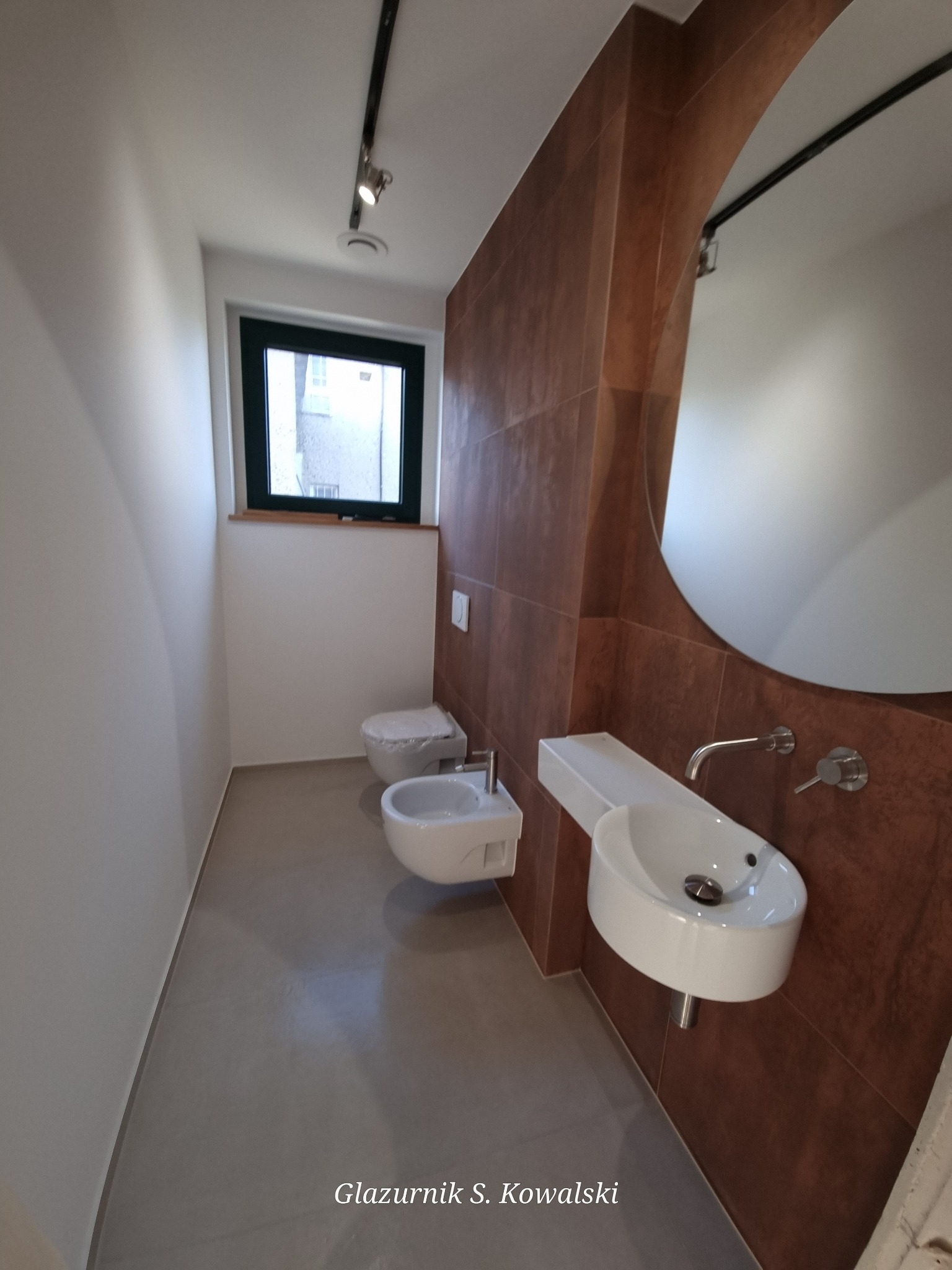 Nowoczesna łazienka z brązowymi płytkami imitującymi drewno, owalnym lustrem z podświetleniem i minimalistyczną białą umywalką oraz toaletą i bidetem w tle.