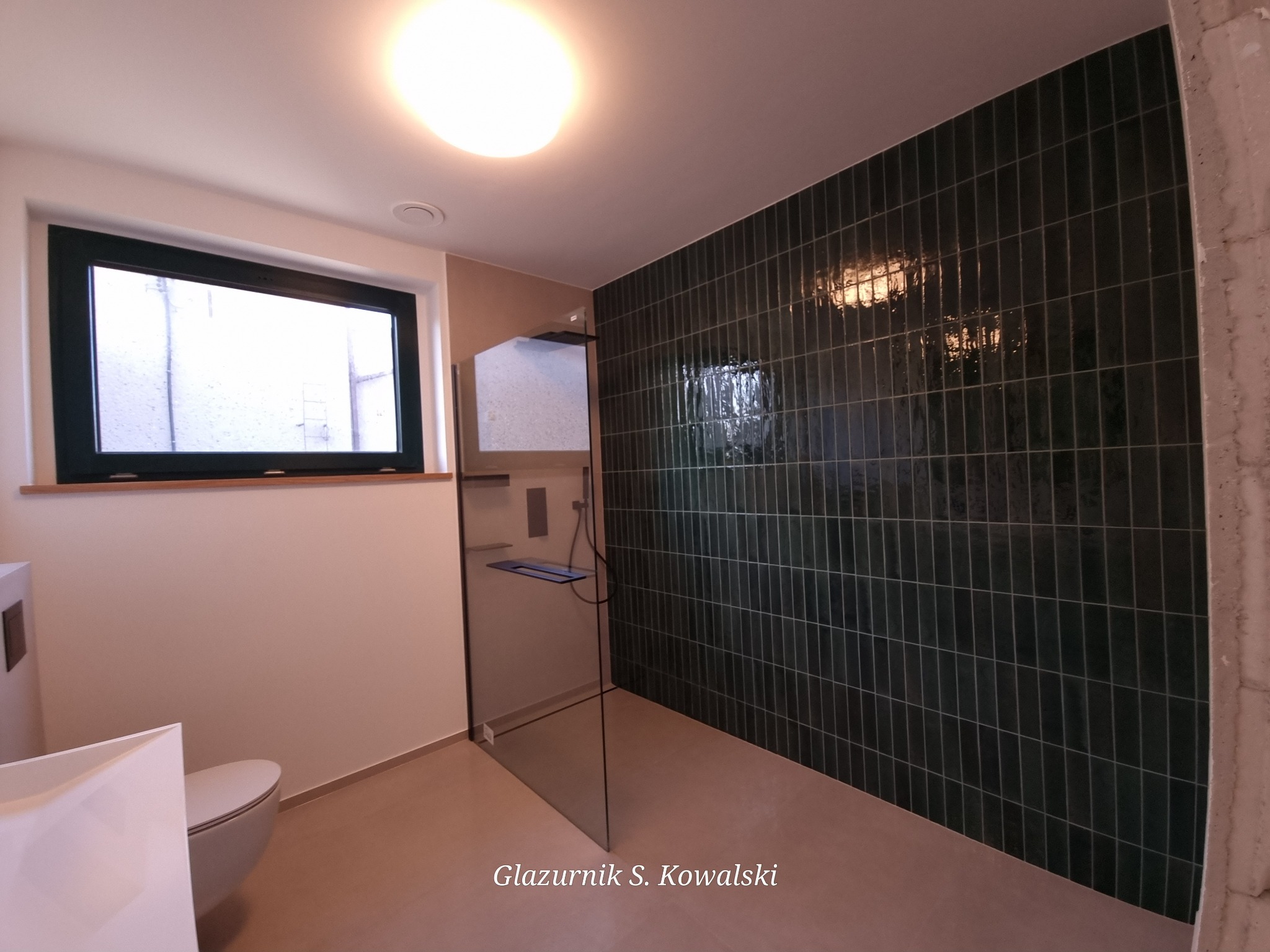 Wykończona łazienka z ciemnozielonymi, pionowo ułożonymi płytkami na ścianie, kabiną prysznicową bez brodzika i wiszącą toaletą. Widoczne surowe wykończenie ściany w rogu.