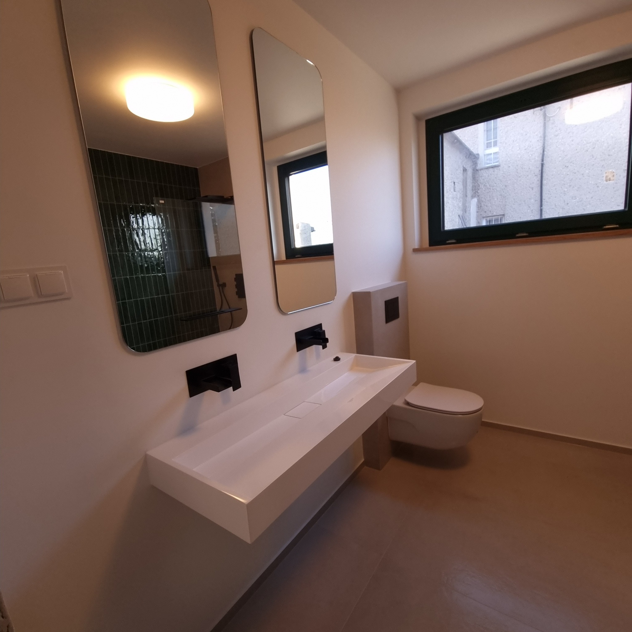 Nowoczesna łazienka z podwieszaną białą umywalką z czarną armaturą, dwoma lustrami o zaokrąglonych rogach, toaletą i oknem z widokiem na budynek z kamienną elewacją.