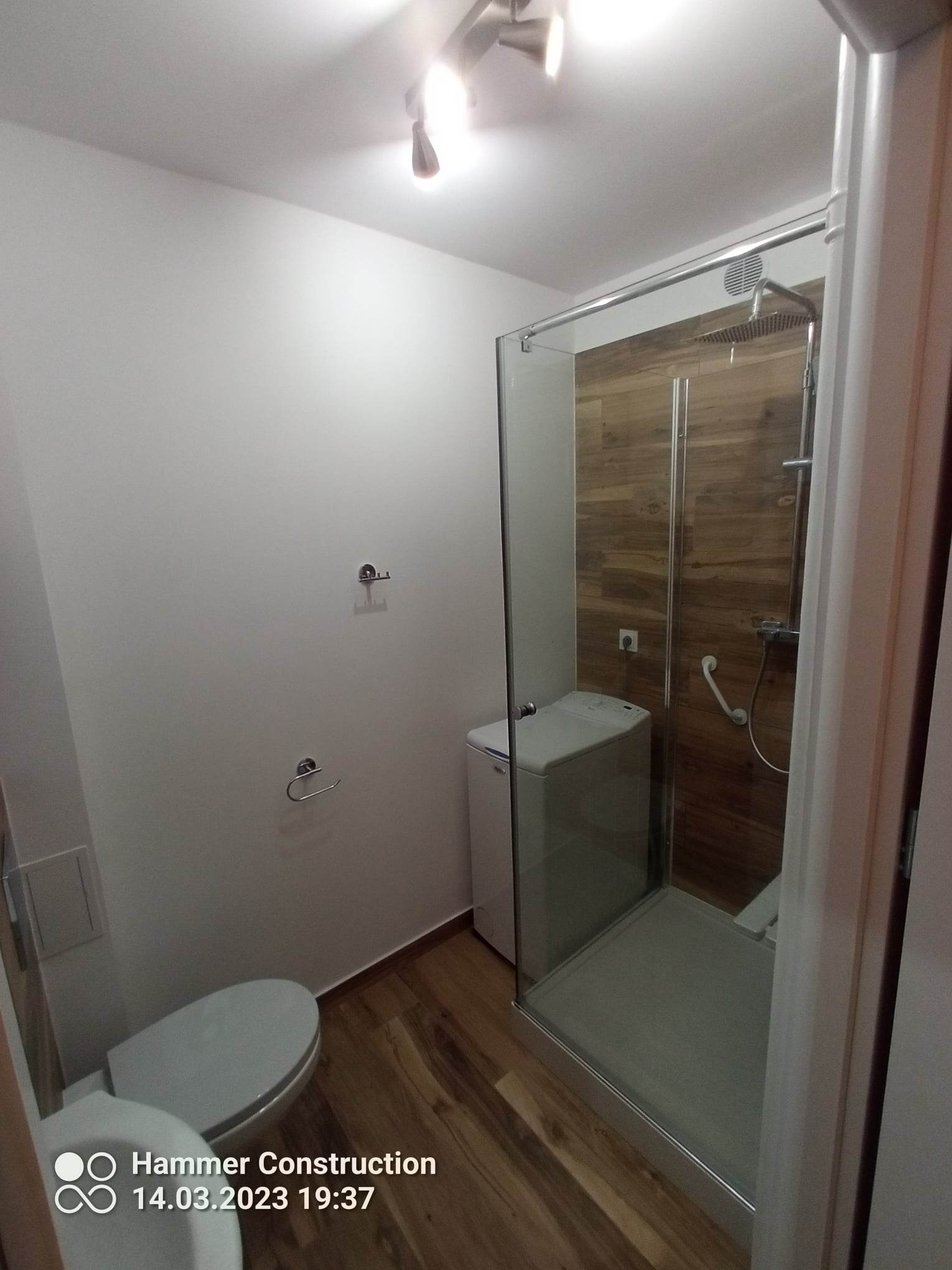 Nowoczesna łazienka z kabiną prysznicową typu walk-in wyłożoną płytkami imitującymi drewno, białą pralką i toaletą, oświetlona trzema punktowymi lampami na suficie.