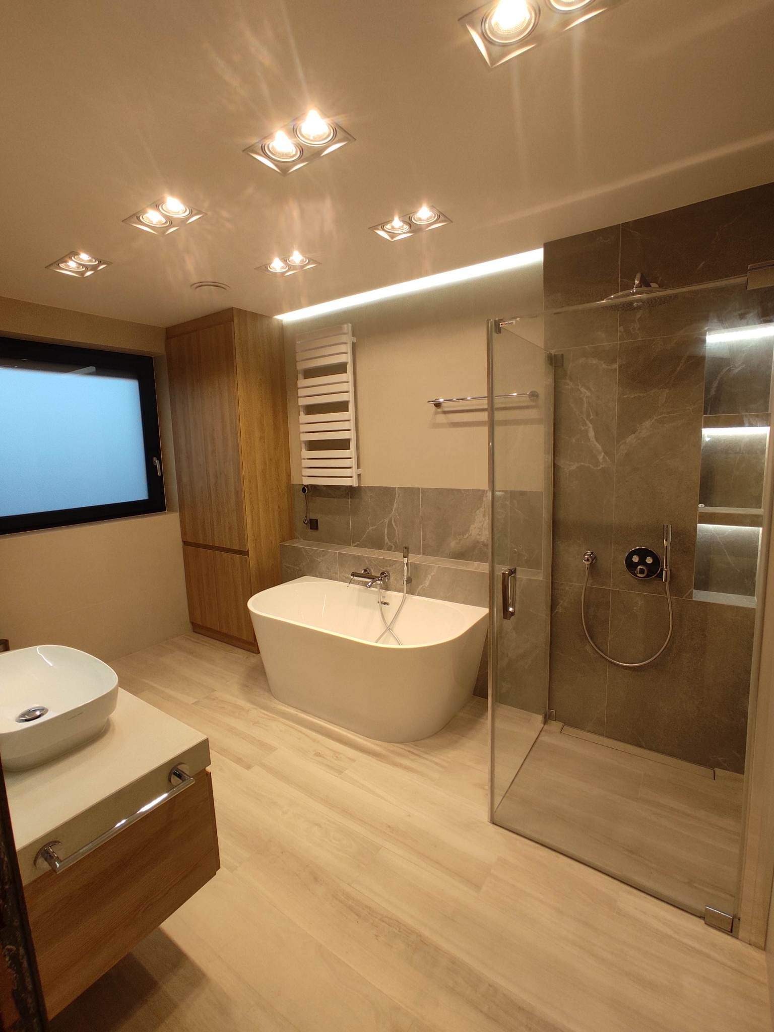 Nowoczesna łazienka z wolnostojącą wanną, kabiną prysznicową typu walk-in z szarymi płytkami imitującymi marmur, drewnianą szafką i umywalką nablatową, oświetlona punktowymi lampami sufitowymi.