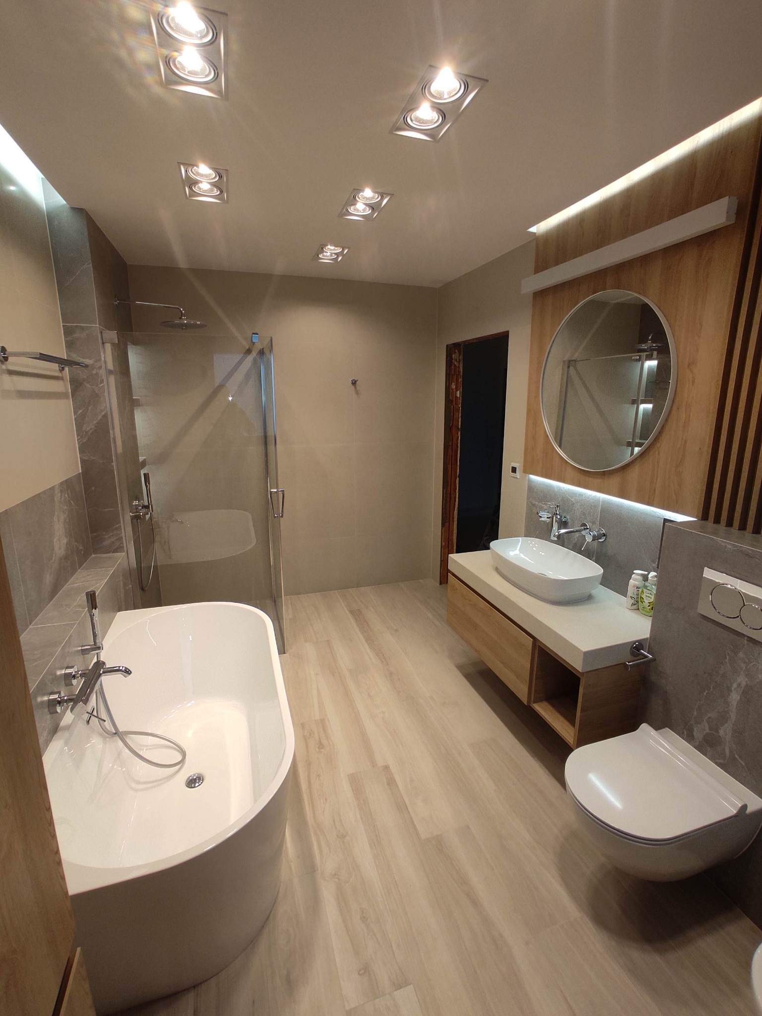 Nowoczesna łazienka z wolnostojącą wanną, kabiną prysznicową typu walk-in, umywalką nablatową na drewnianej szafce i podwieszaną toaletą, wykończona płytkami imitującymi drewno i szarym marmurem.