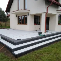 Nowy taras z kostki brukowej w Kościerzynie, z trzema schodami obramowanymi ciemną kostką, prowadzący do jasnego domu z beżową elewacją, widoczny z boku.