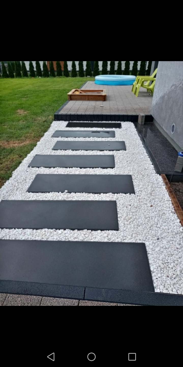 Ścieżka z prostokątnych, grafitowych płyt ułożonych w białym żwirze, prowadząca od tarasu z piaskownicą i basenem do ściany domu.