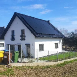 Biały dom jednorodzinny z czarną dachówką i panelami fotowoltaicznymi na dachu, widoczny zza ogrodzenia z siatki, z trzema kolorowymi pojemnikami na śmieci po lewej stronie.