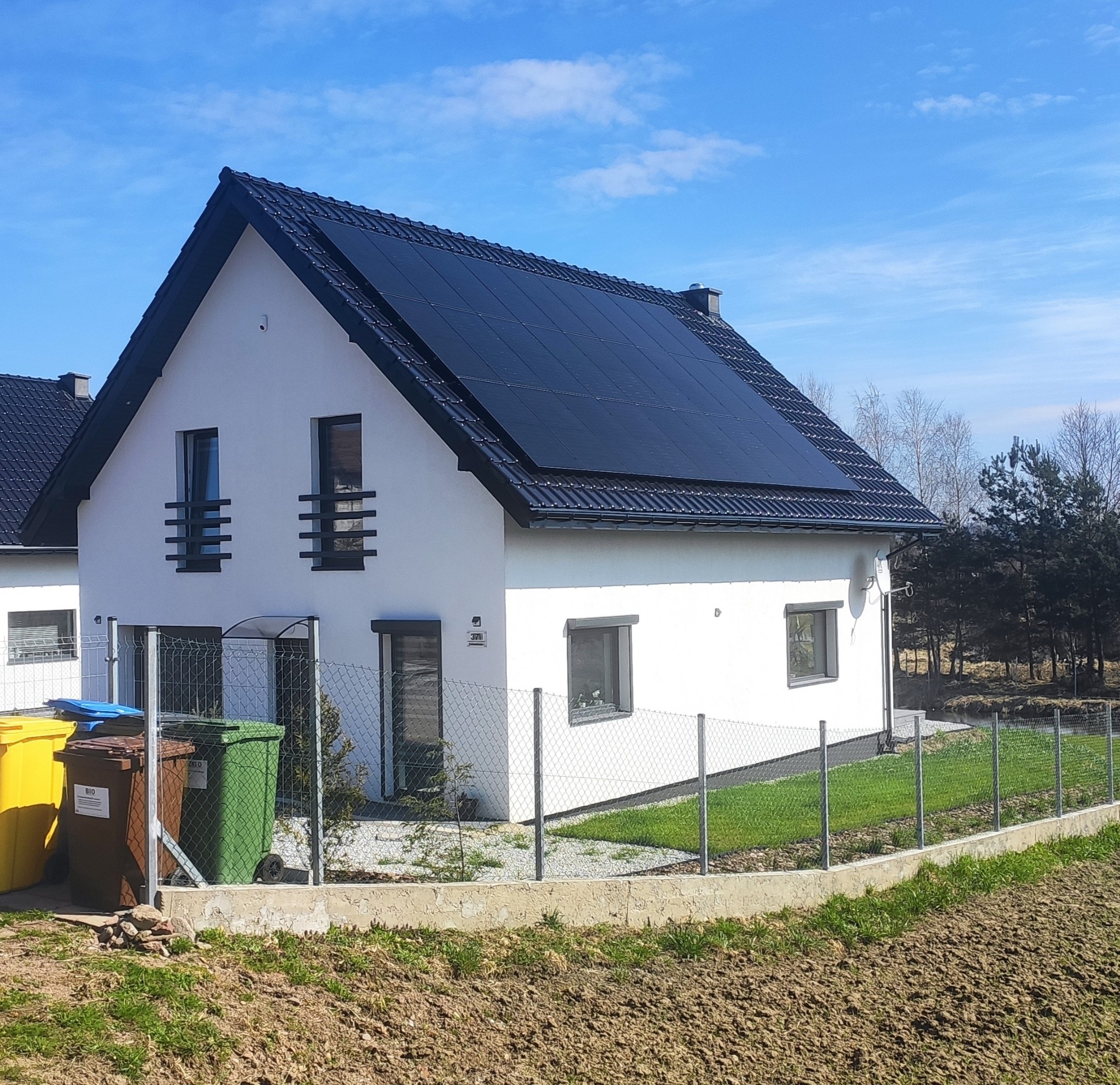 Biały dom jednorodzinny z czarną dachówką i panelami fotowoltaicznymi na dachu, widoczny zza ogrodzenia z siatki, z trzema kolorowymi pojemnikami na śmieci po lewej stronie.