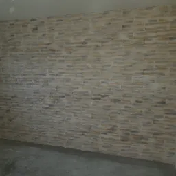 Ściana wyłożona dekoracyjnym kamieniem w odcieniach beżu i szarości, widoczny surowy beton na podłodze, pomieszczenie w trakcie wykańczania.